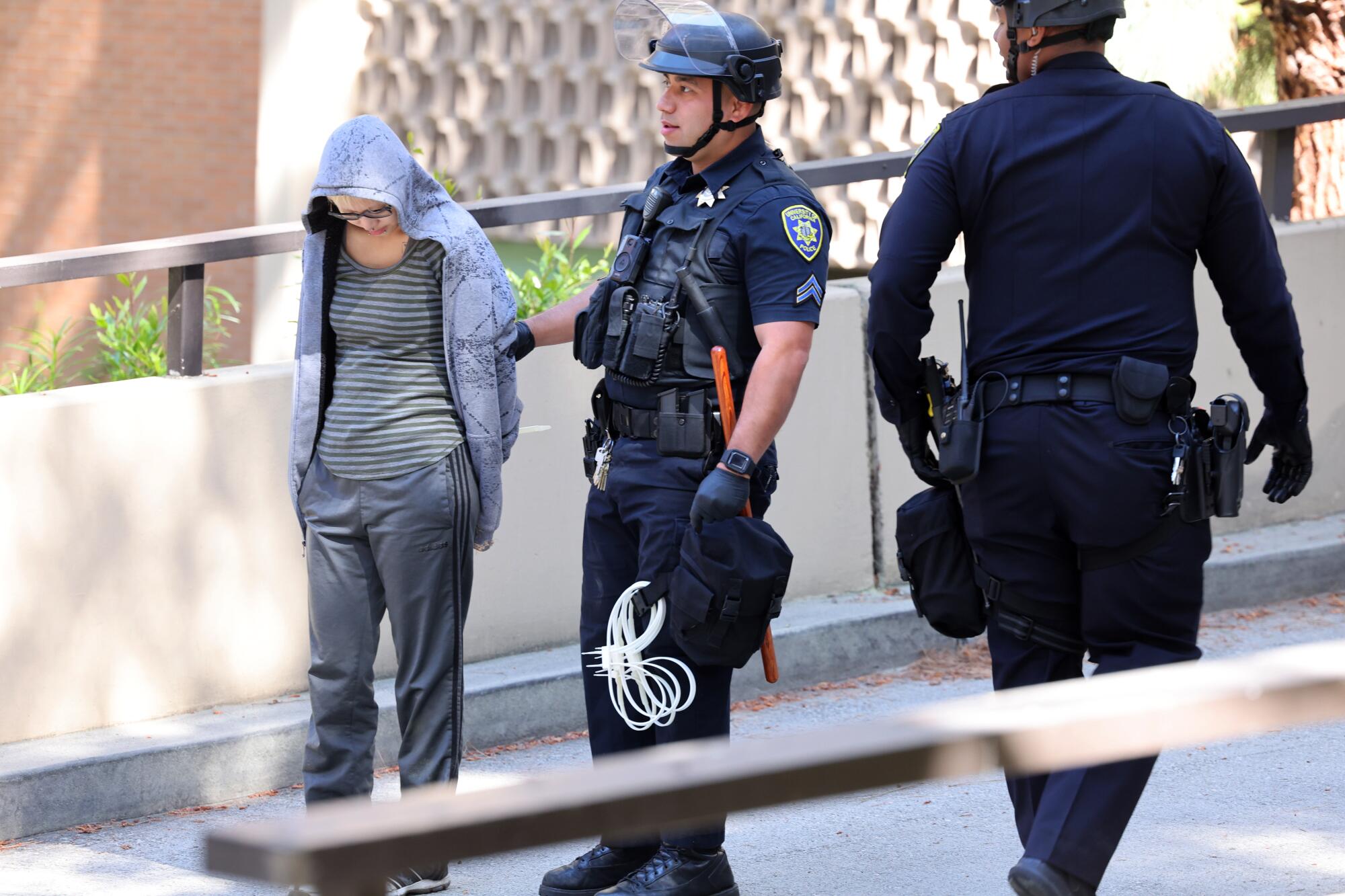 Police stand next to a person in a hoodie and handcuffs.