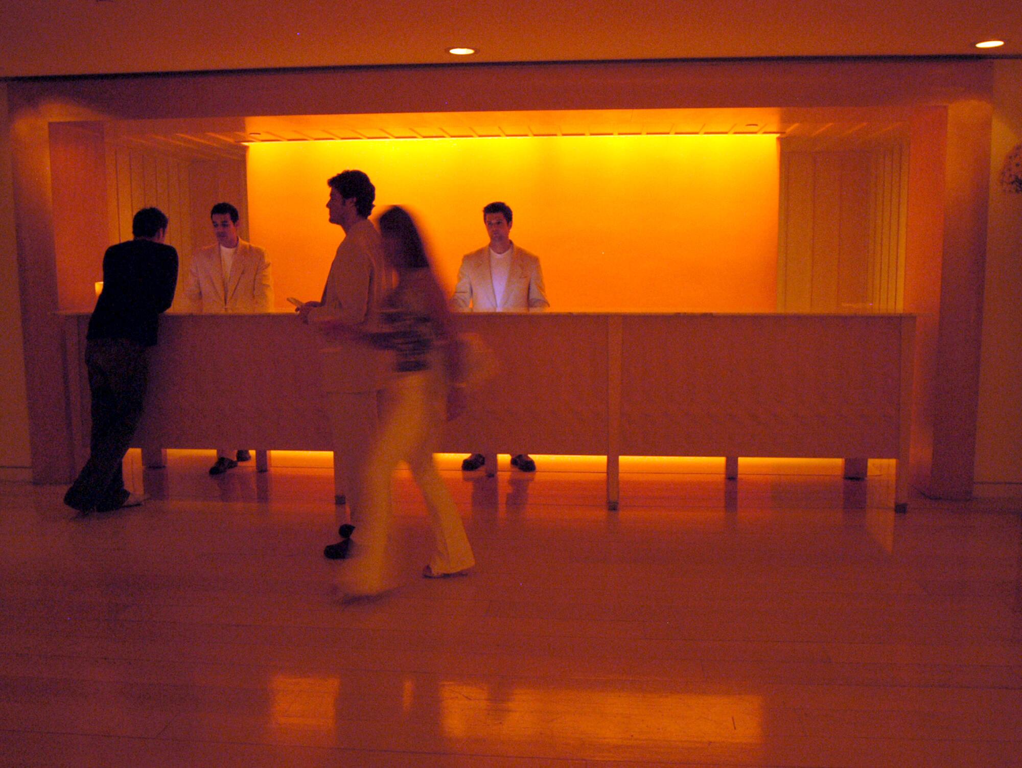 The lobby of the Mondrian Hotel in a moody orange hue. Two staff members stand behind a long desk while guests pass by.
