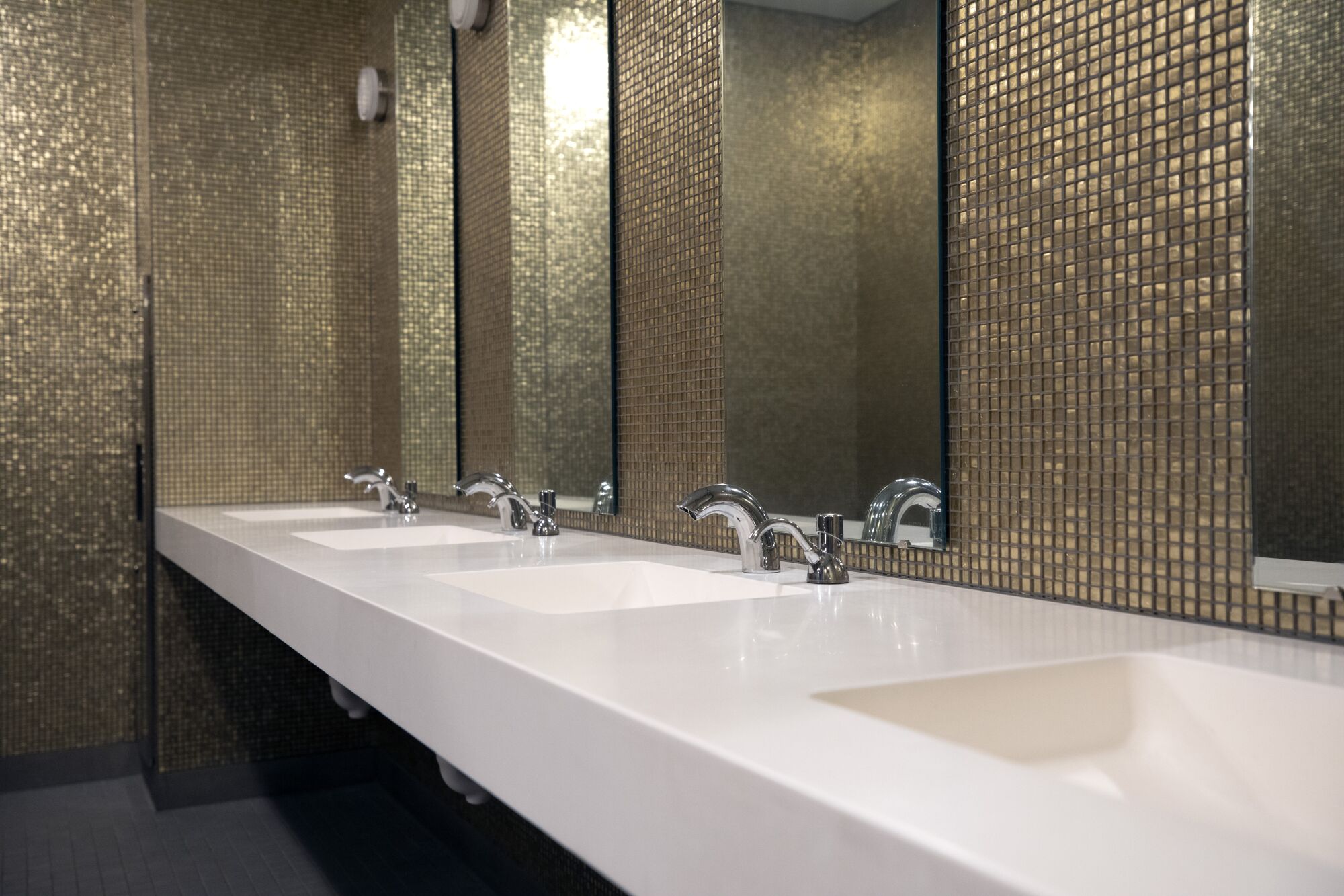 A horizontal view of a bathroom shows a row of white sinks in front of a wall of golden mosaic tile.