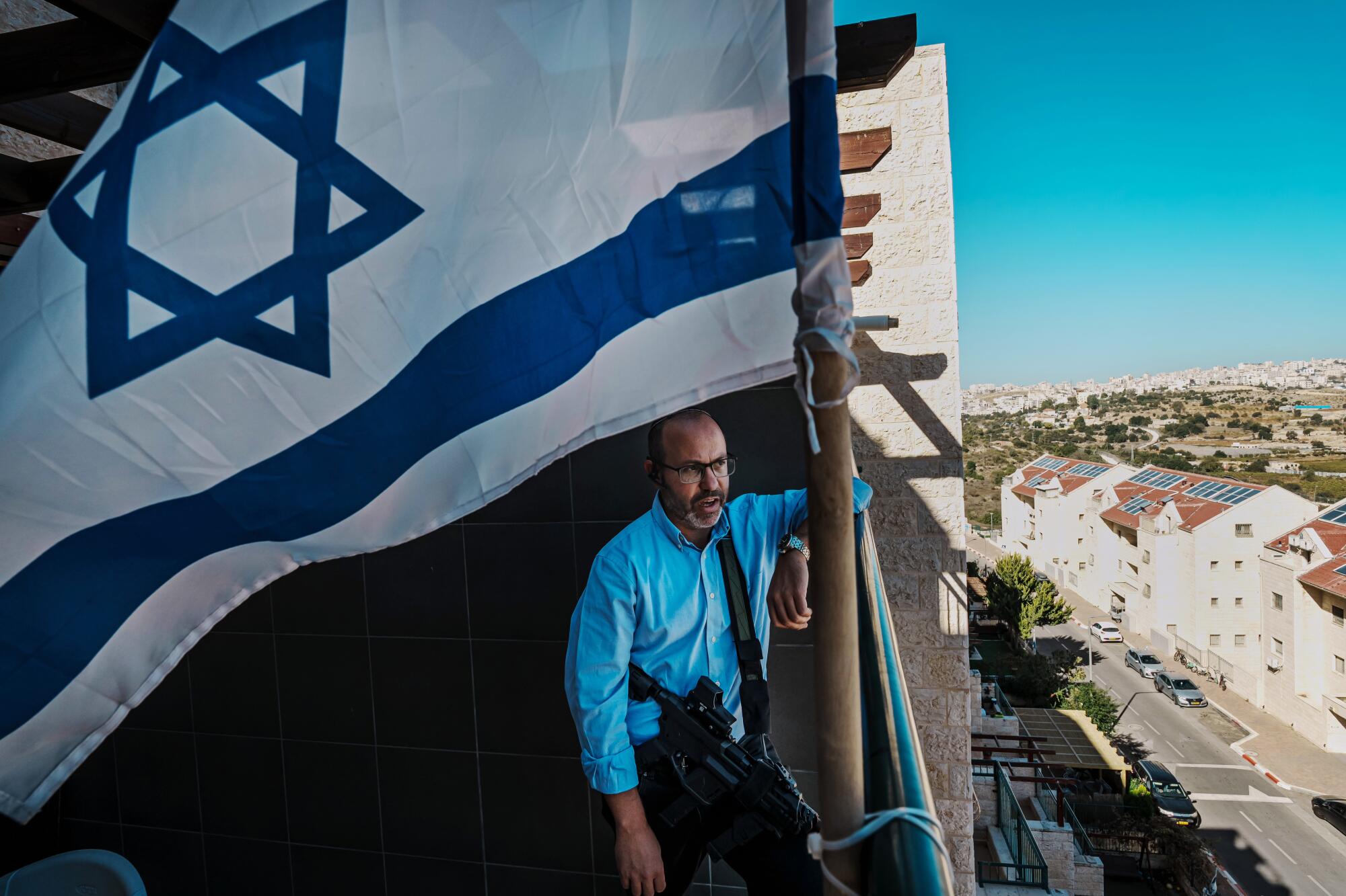 A man in a blue shirt, with an assault weapon slung over his waist, stands near a large white-and-blue flag with a blue star