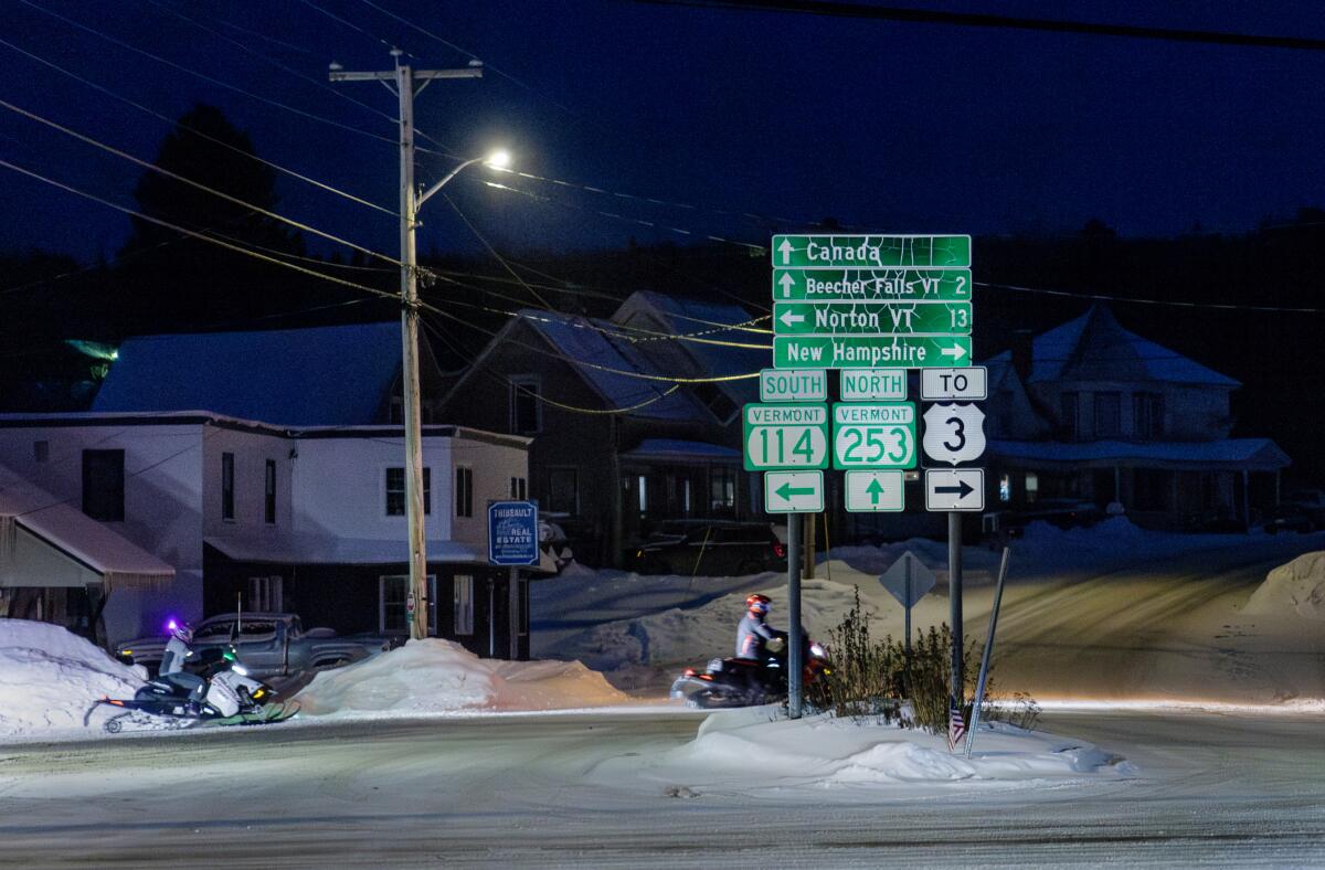 Snowmobilers drive past signage that points the direction to New Hampshire, Vermont and Canada.