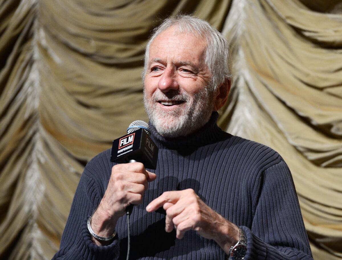 A head-and-shoulders frame of a man with grey hair and beard, smiling while holding a microphone