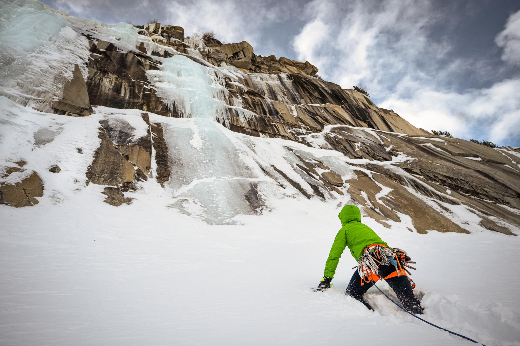A person wearing a green jacket crouches below an iced-over cliffside.  