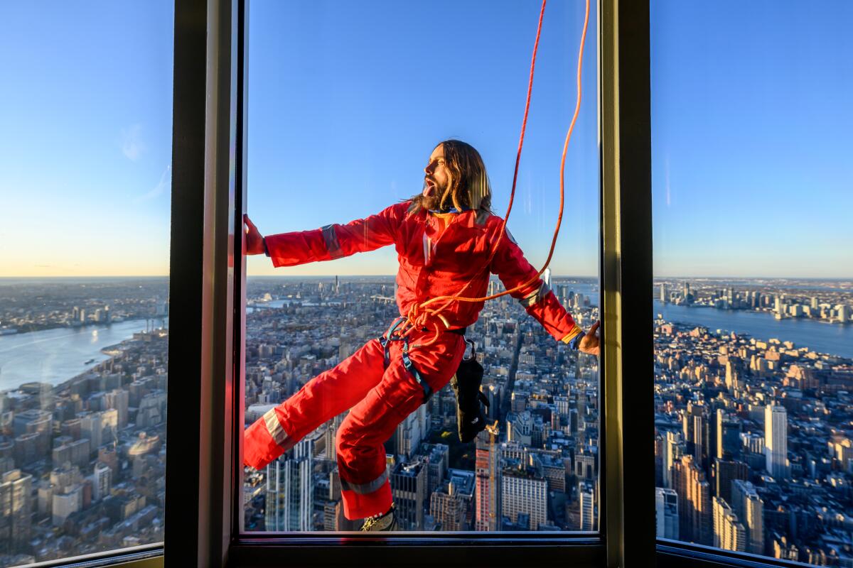 Jared Leto is seen through a window climbing the Empire State Building in an orange jumpsuit with Manhattan behind him.