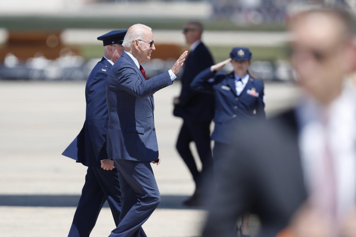 President Biden waves as he walks with a uniformed military officer and another military member salutes 