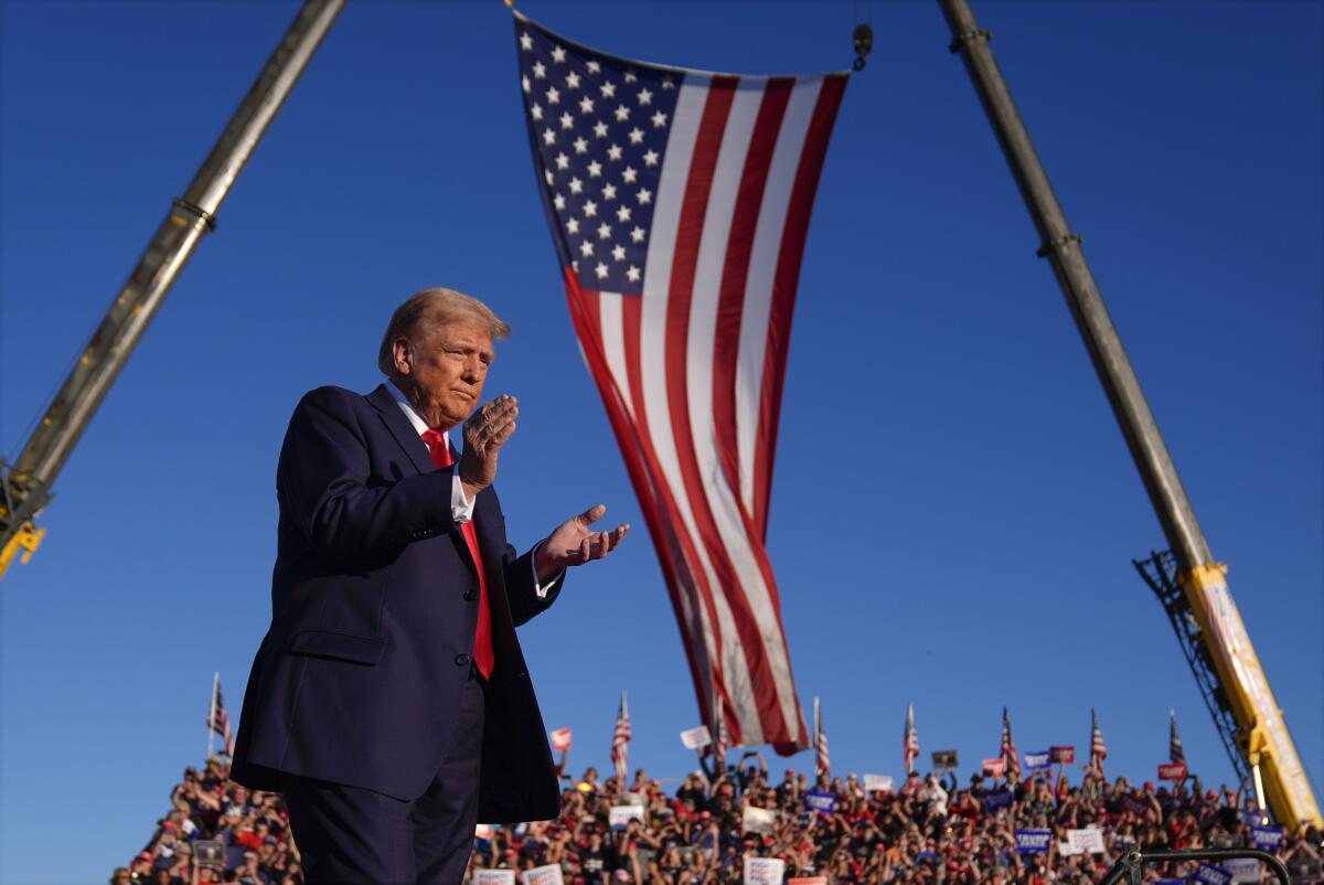 Former President Trump claps with a crowd and large American flag in the background.