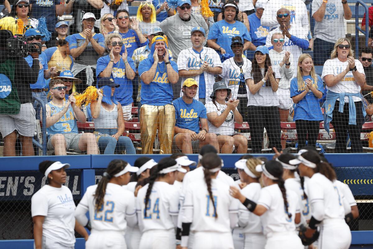 UCLA softball fans cheer in the stands as players huddle on the field.