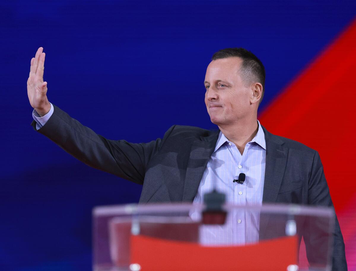 Richard Grenell stands behind a see-through lectern and against a split red and blue backdrop, waving