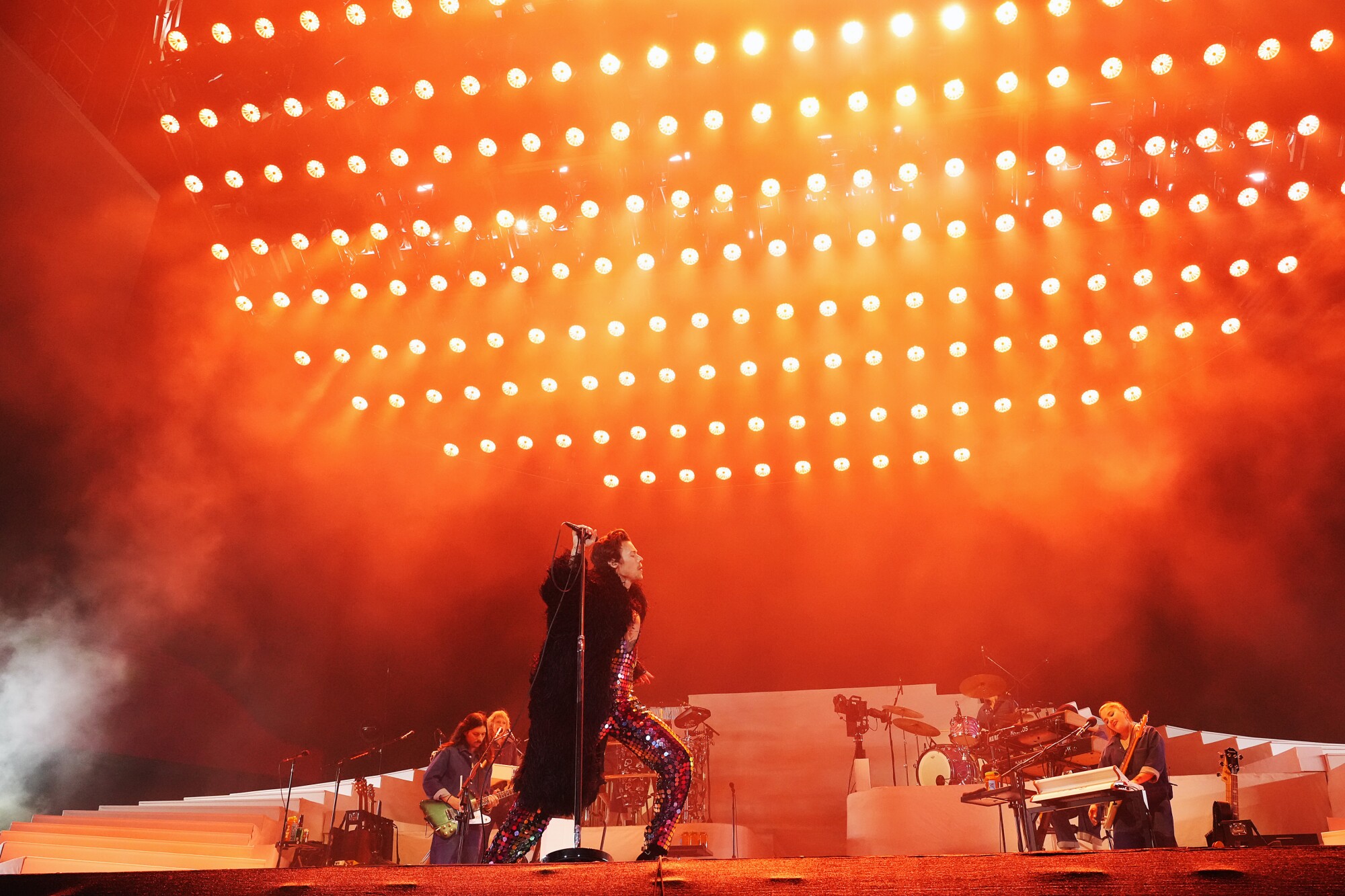 Harry Styles walks onstage in a coat and sparkly jumpsuit while rows of lights shine through smoke above him.