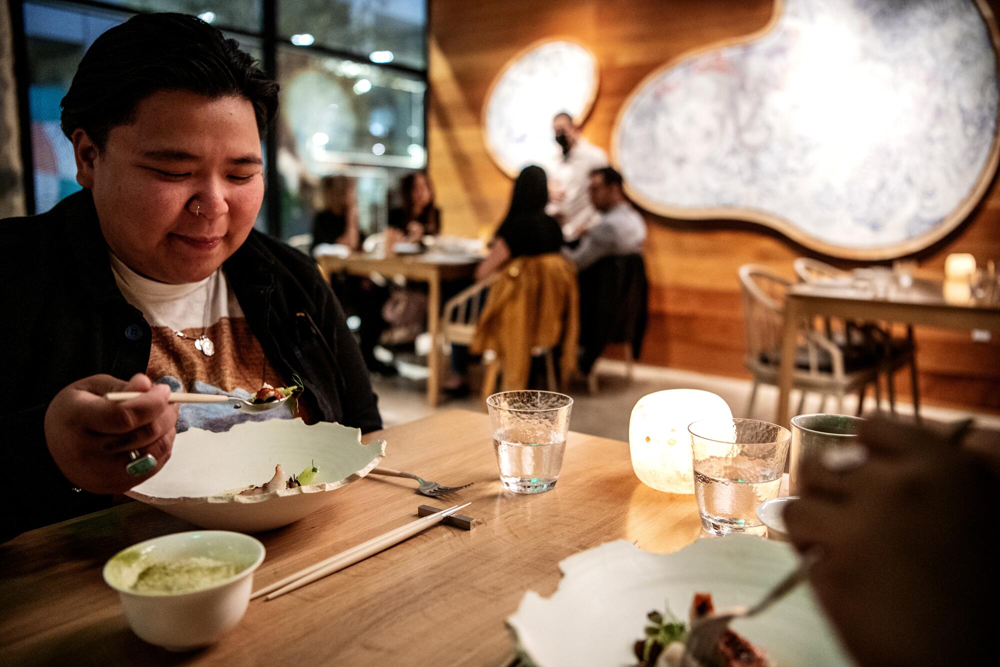 A person eats at a wooden table, with other diners in the background, at Kato in L.A.
