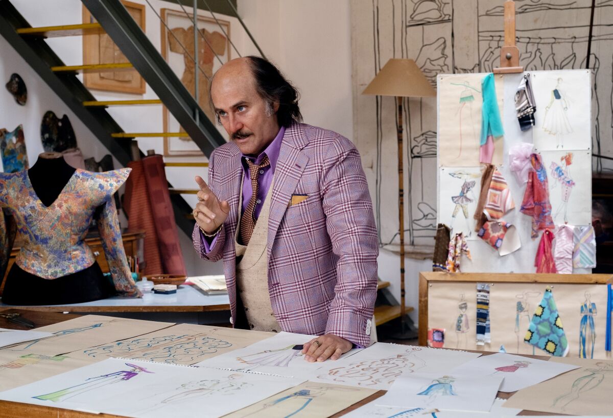 A balding man in a purple plaid suit stands in front of a table full of fashion sketches.