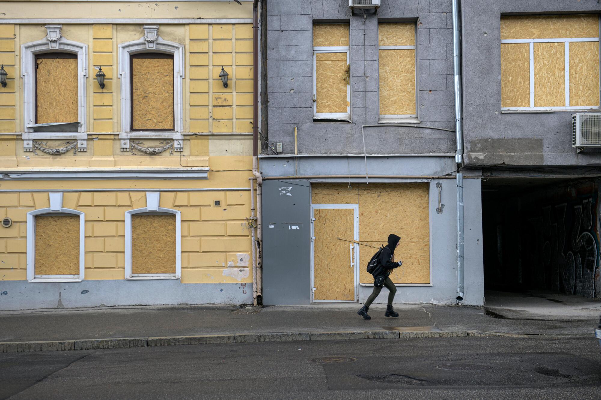 A person passes buildings with boarded-up windows and doors.