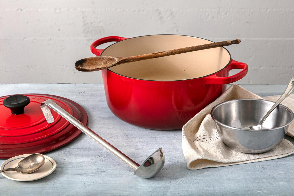 A red Dutch oven, with a wooden spoon across the top, and lid with a ladle, two spoons, a dish towel and a silver bowl.