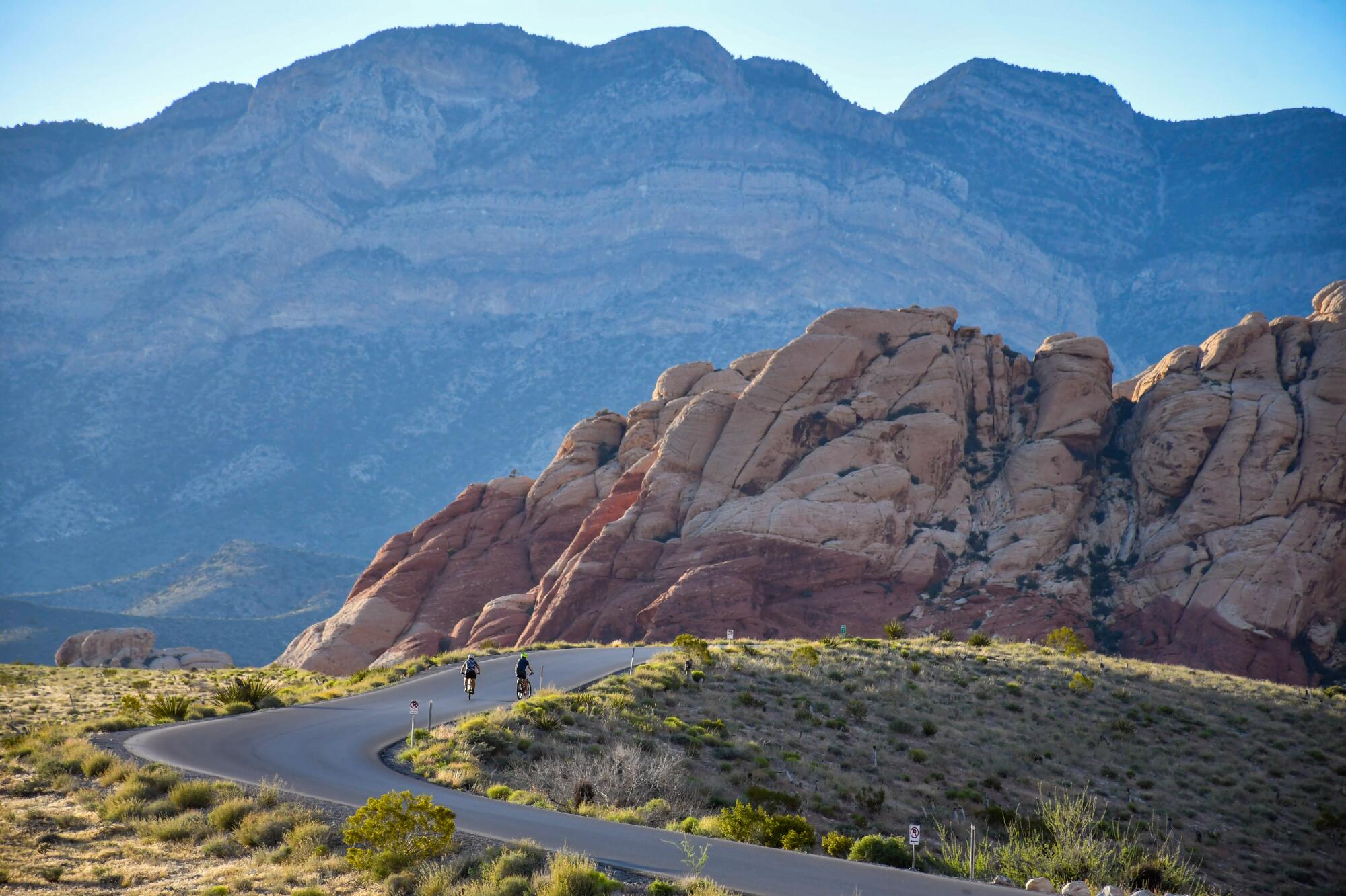 People bicycle on a road toward red rock hills, with larger hills in the distance