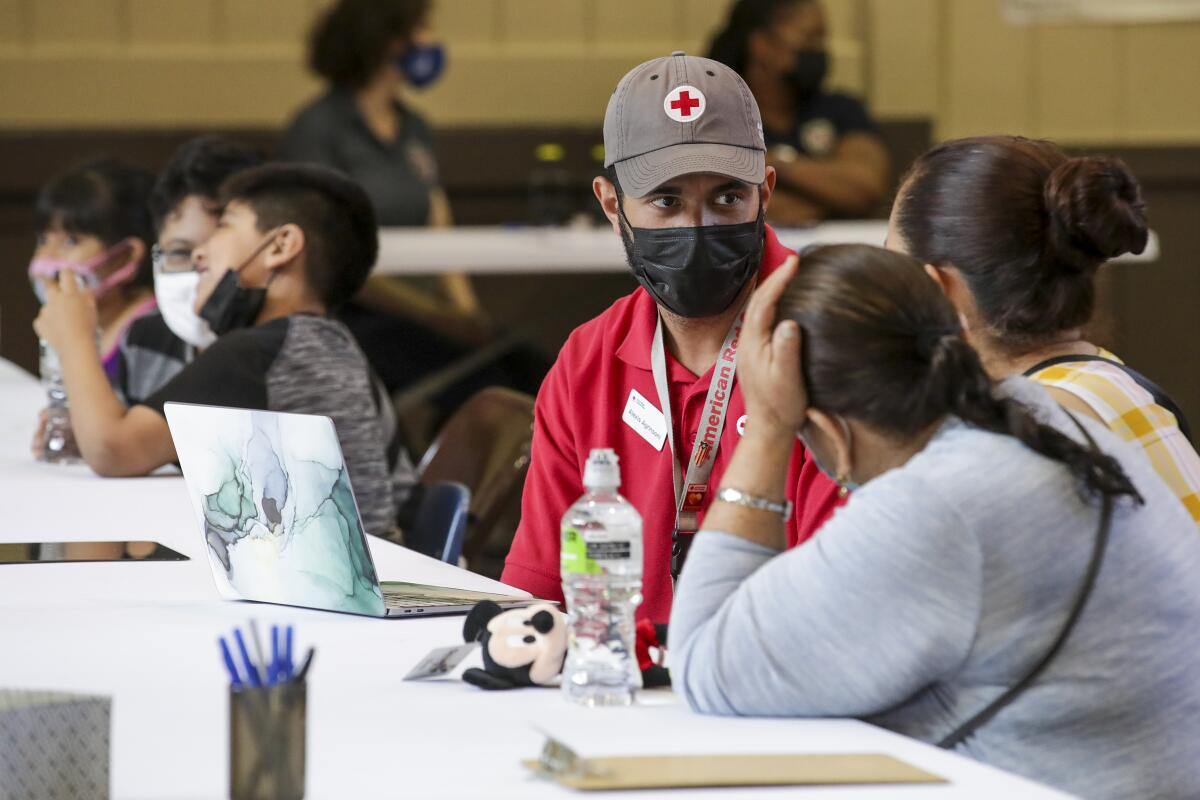A man in a cap with a red cross on it speaks to two women at a long table.