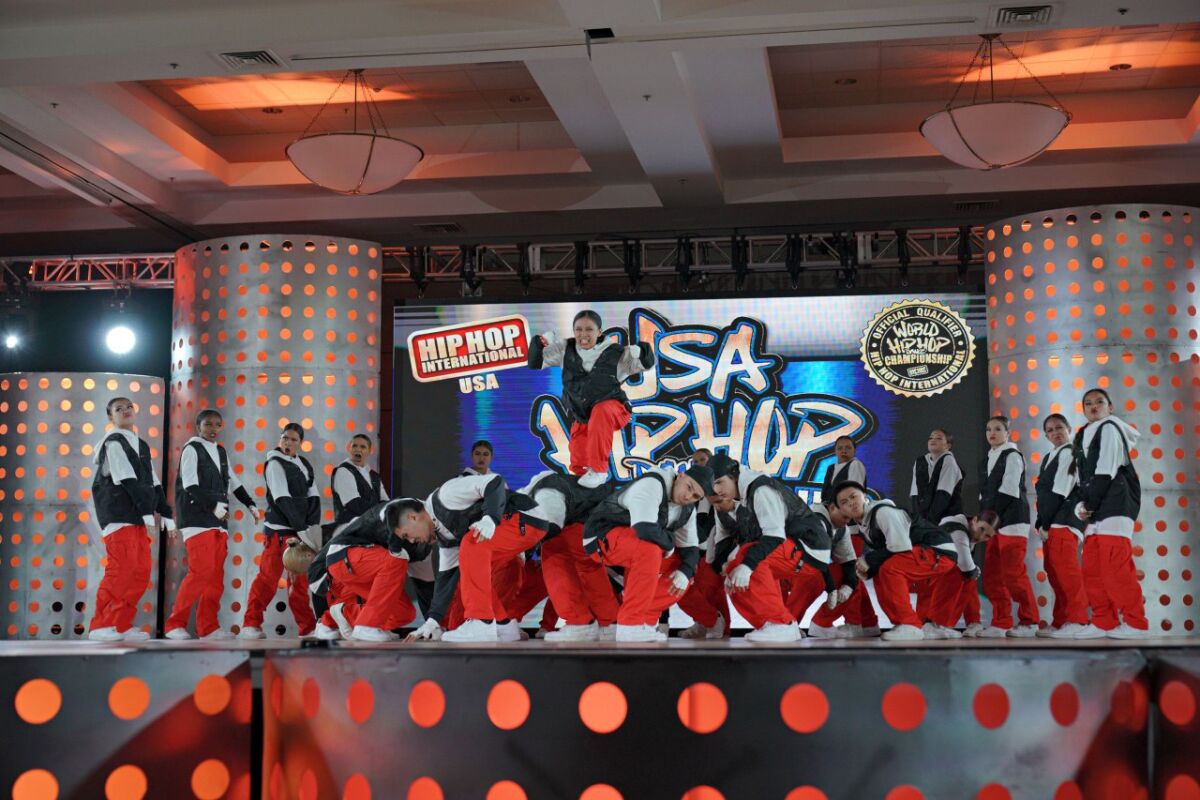 A hip-hop dance crew in red pants and black-and-white shirts performs onstage.