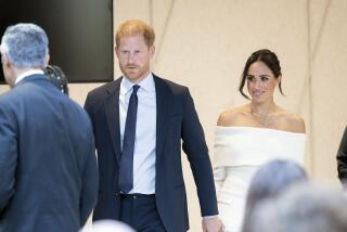 Prince Harry in a suit and Meghan in a white dress hold hands as they walk through a crowd