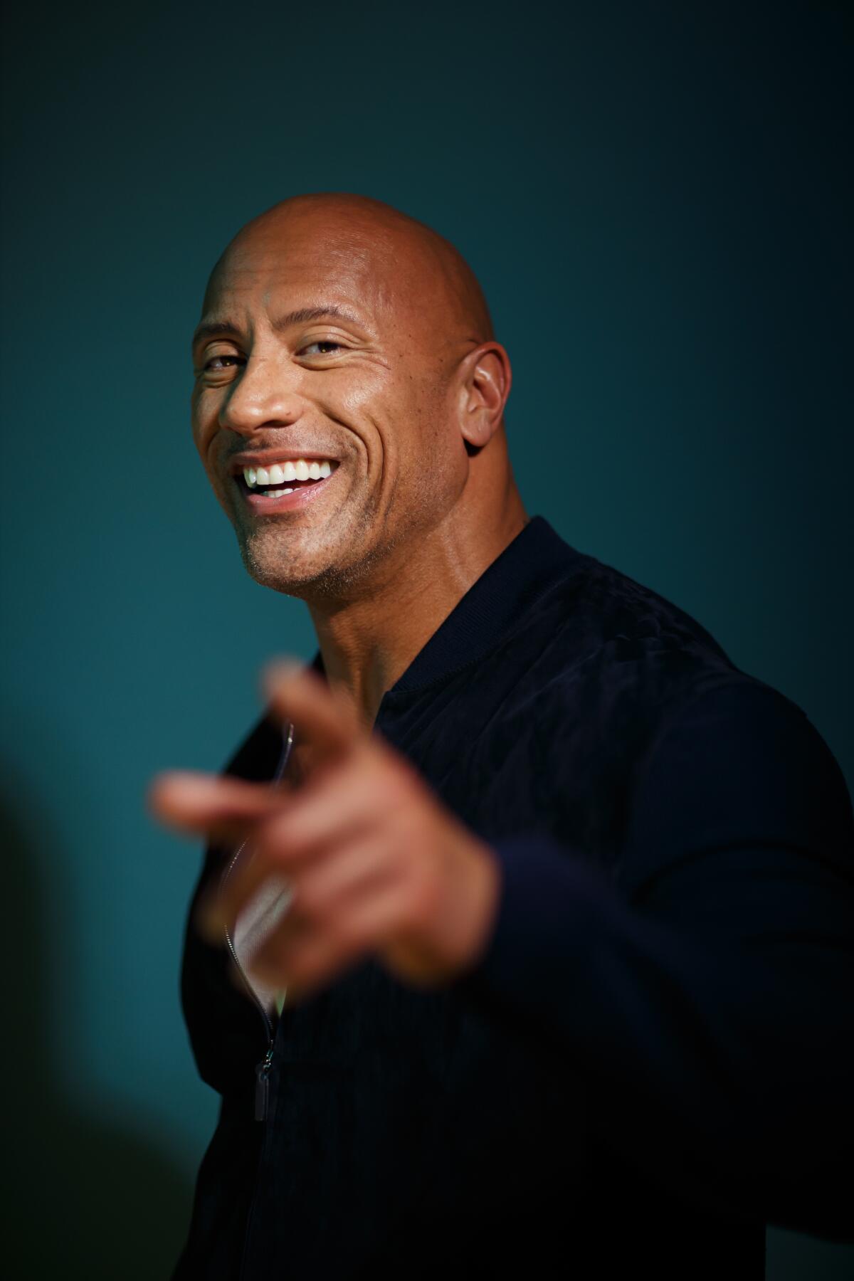 Dwayne Johnson smiles and points with one hand while wearing a black long sleeve shirt