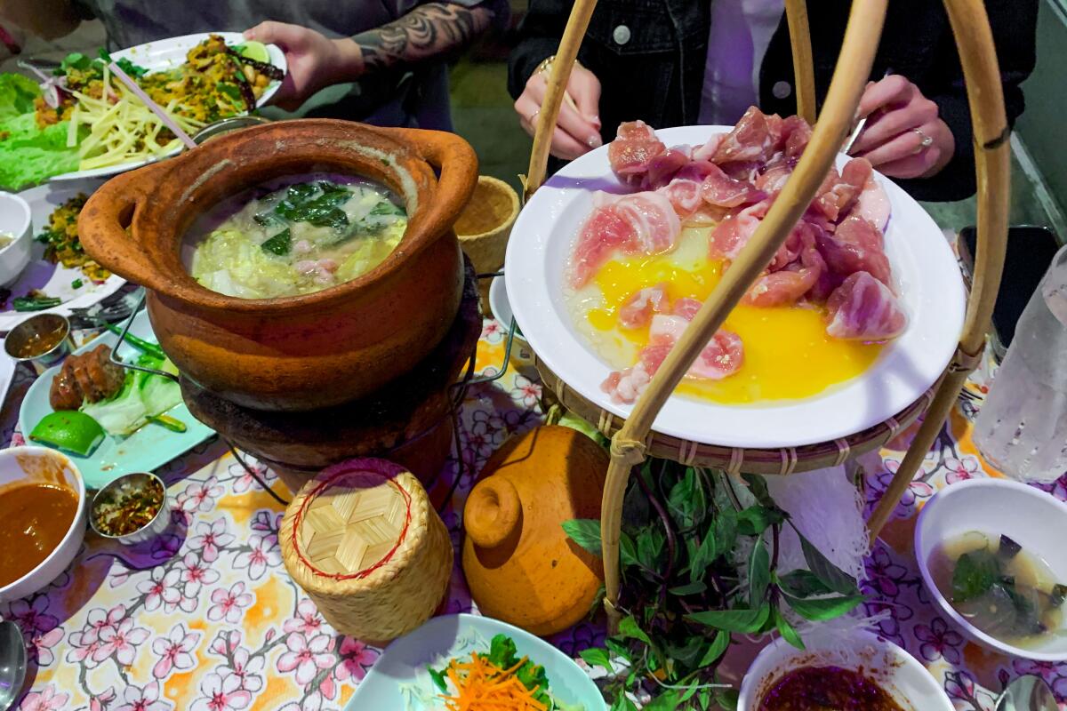 A clay pot of soup on a table next to a tower with raw meats for cooking