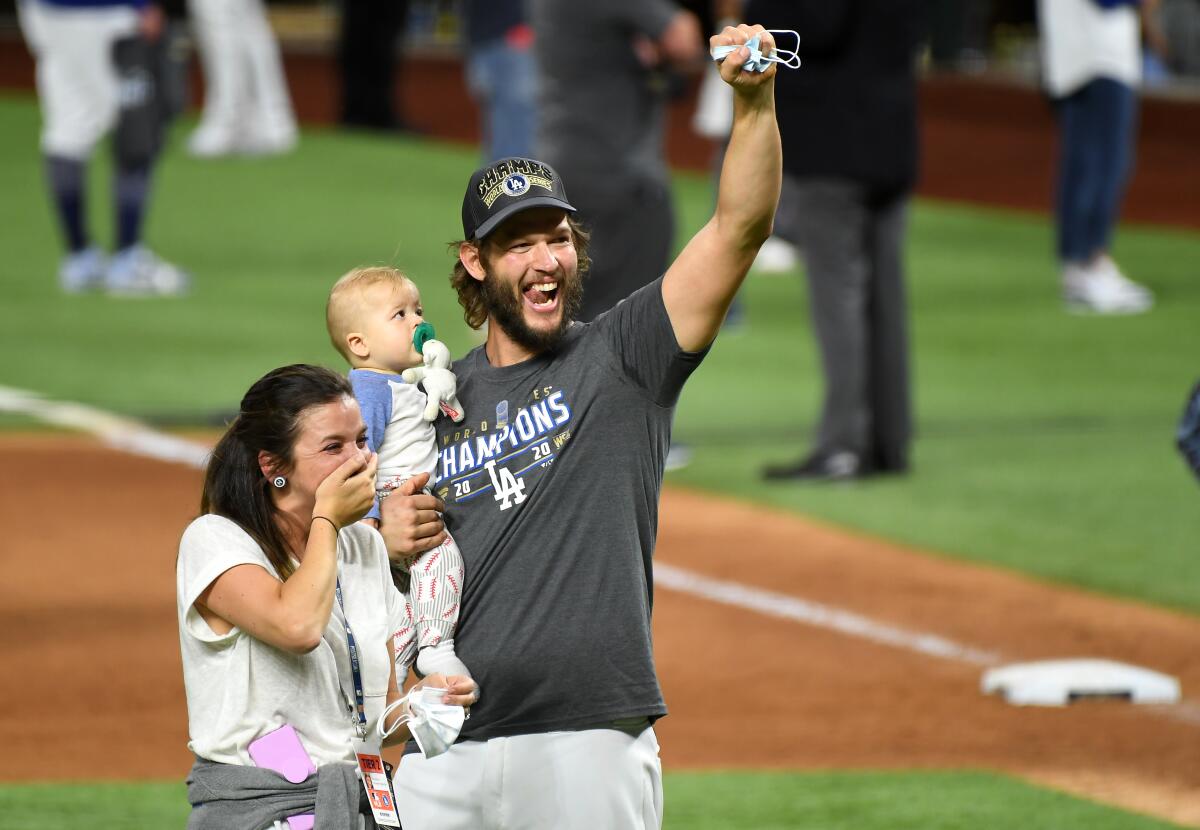 A smiling Clayton Kershaw, holding his baby, stands with his wife on the field and raises a fist.