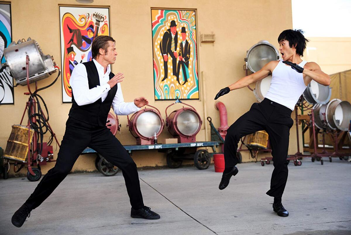 A man in a tux vest stands in a fighting position toward another man preparing to karate kick at him 