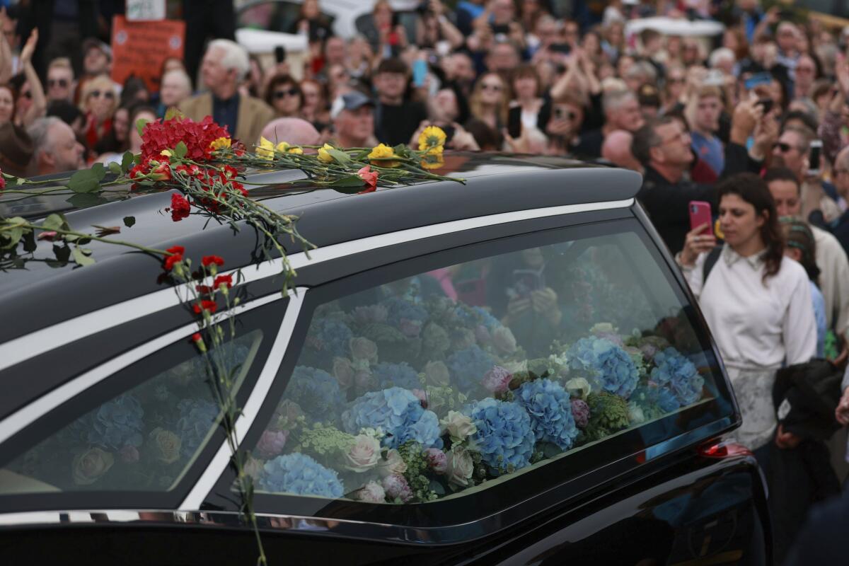 A hearse filled with blue and pink flowers with red and yellow flowers on top in front of a crowd of people