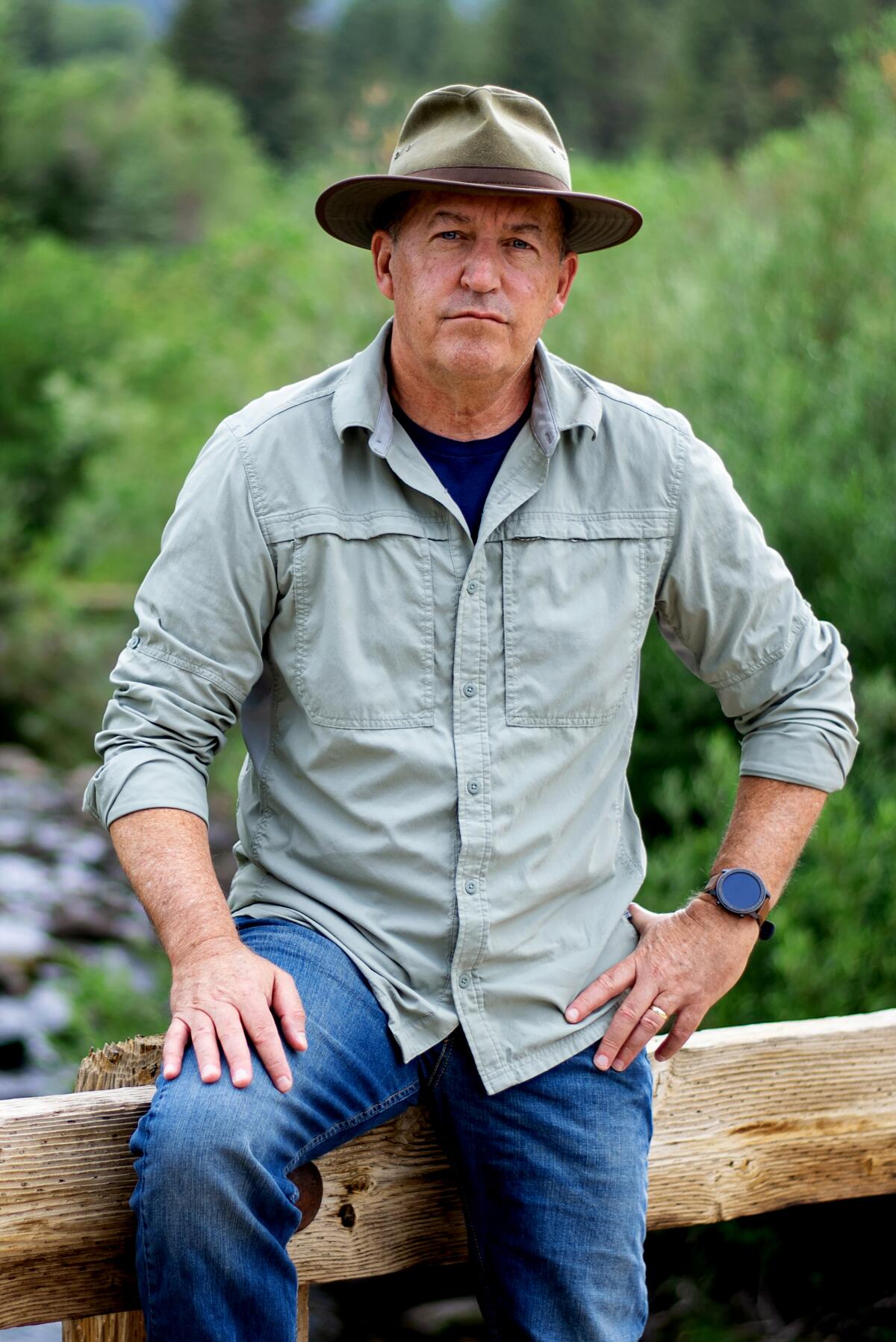 A man wearing jeans and a hat poses against a fence, with shrubbery in the background.