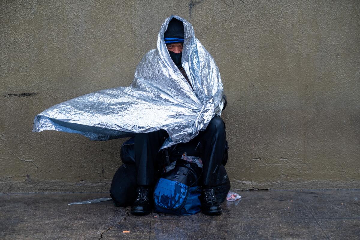 A man sits on the sidewalk wrapped in a foil-like blanket whipped by the wind