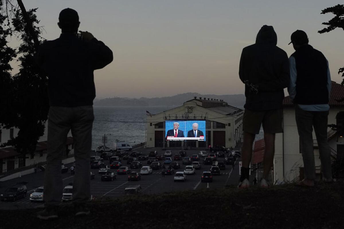 At dusk, people are seen in silhouette. Below cars are parked in a lot in front of a large screen.
