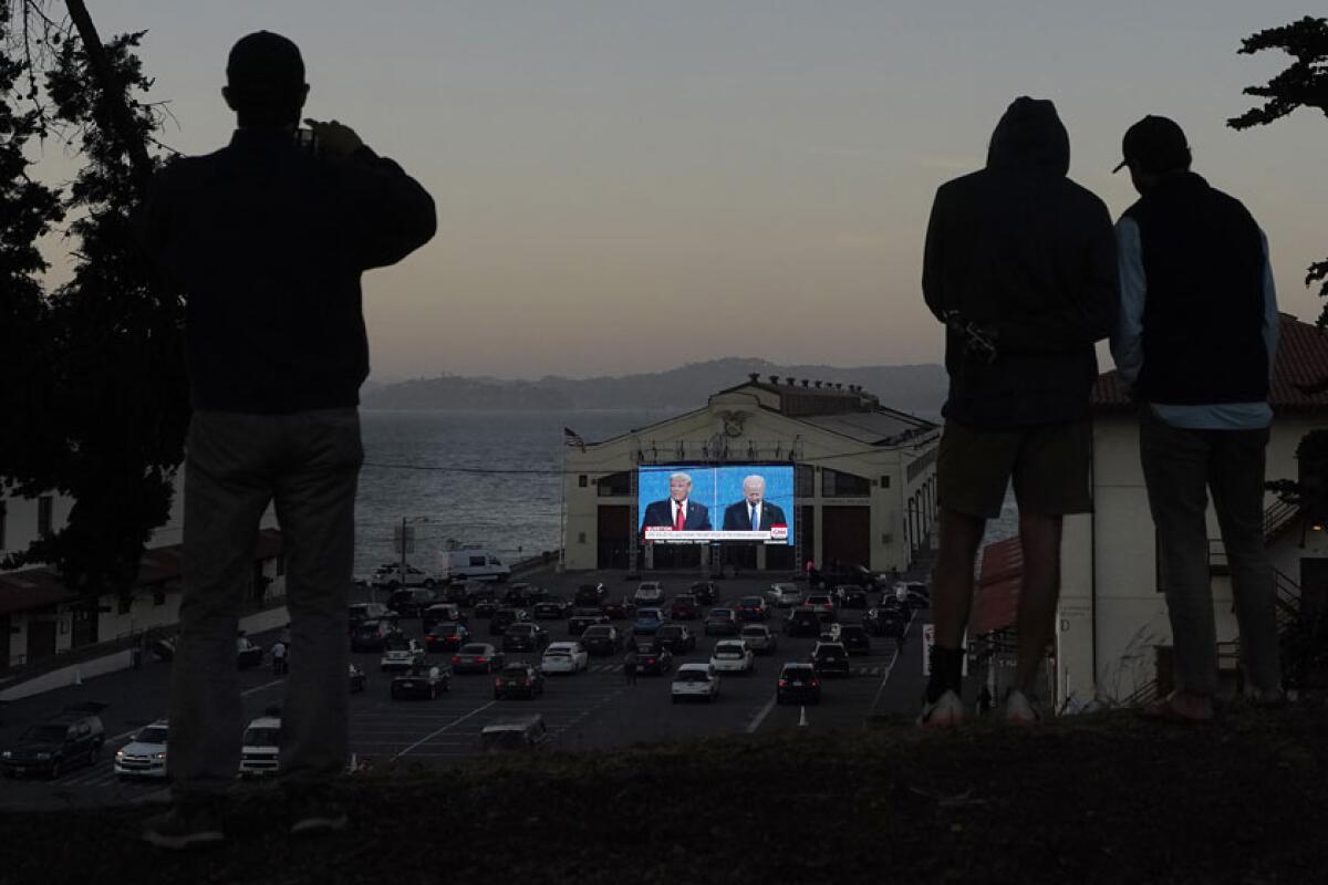 At dusk, people are seen in silhouette. Below cars are parked in a lot in front of a large screen.