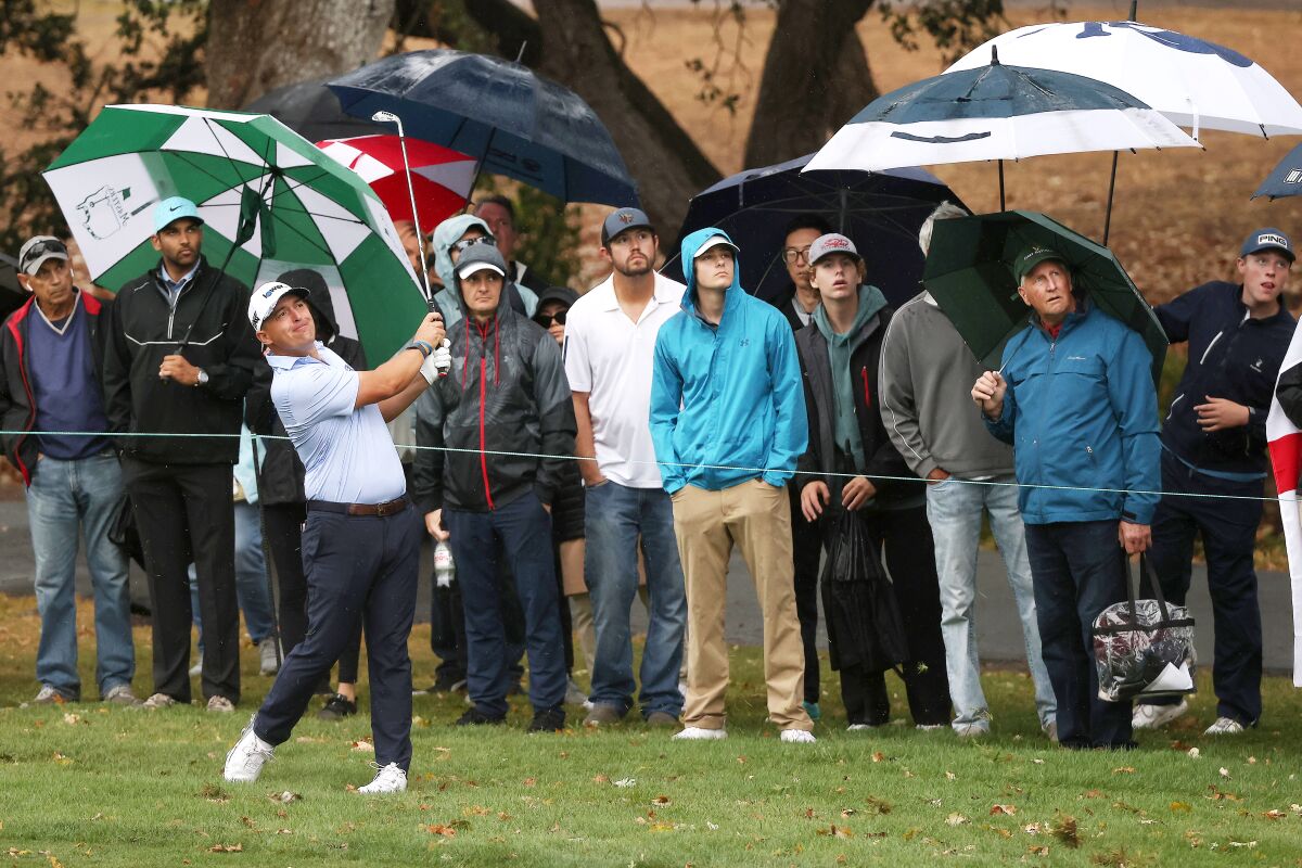 A golfer swings a club while spectators watch in the rain.