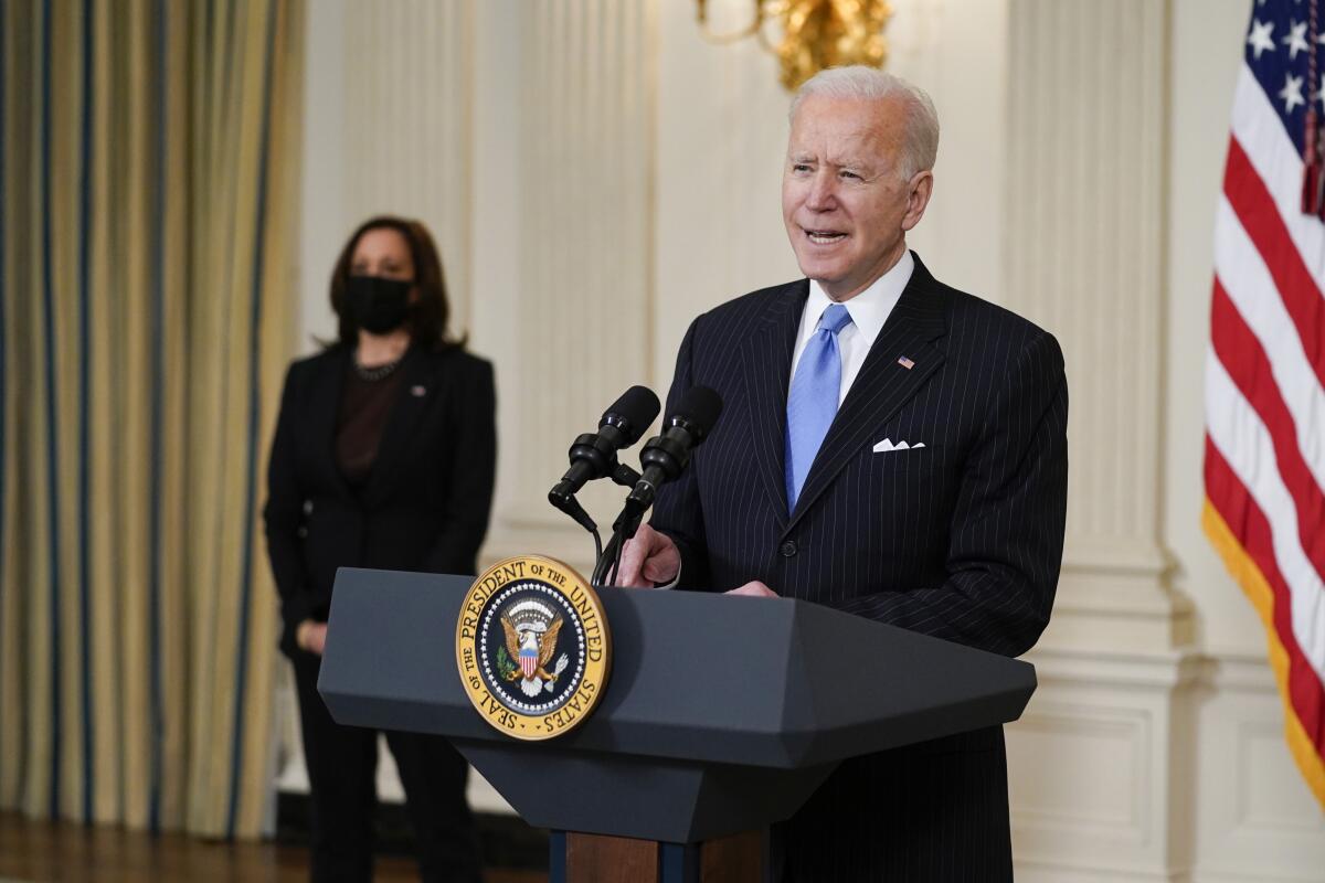 Biden speaks at a podium. Harris wears a mask and stands behind him.