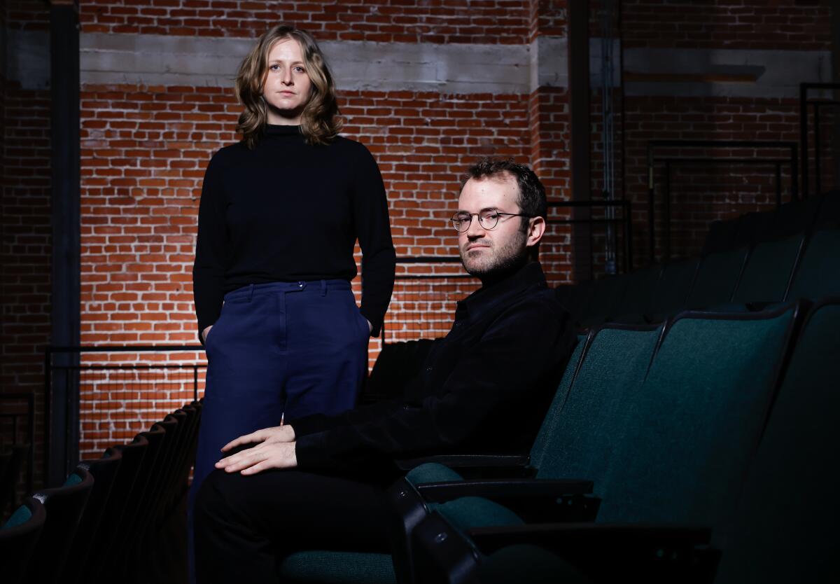 Micah Gottlieb, right, sits in a chair, and Sarah Winshall, stands to his left in front of a brick wall.