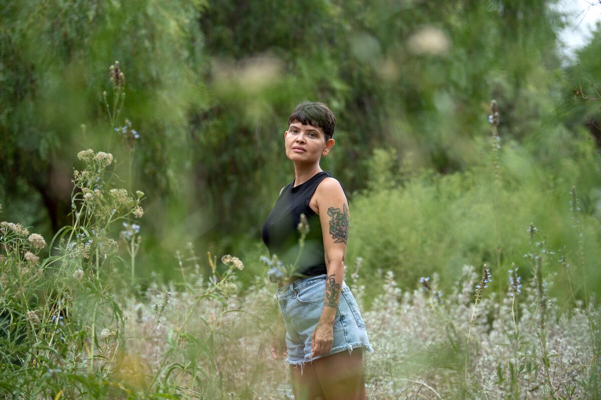 A woman with tattoos in a tank top and cut-off denim shorts and a short haircut in a meadow of wildflowers.