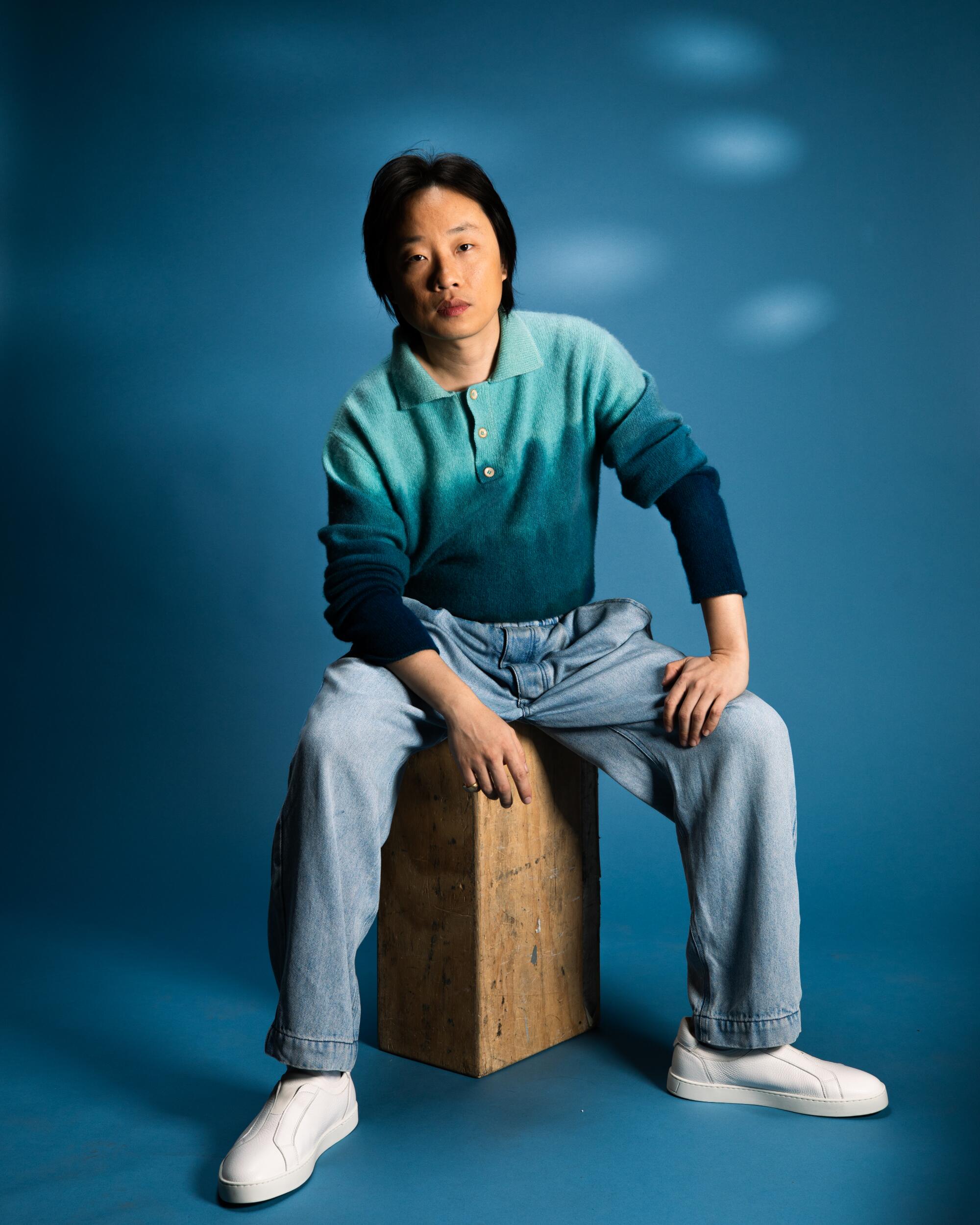 Jimmy O. Yang in a blue long-sleeve shirt and jeans sitting on a box.