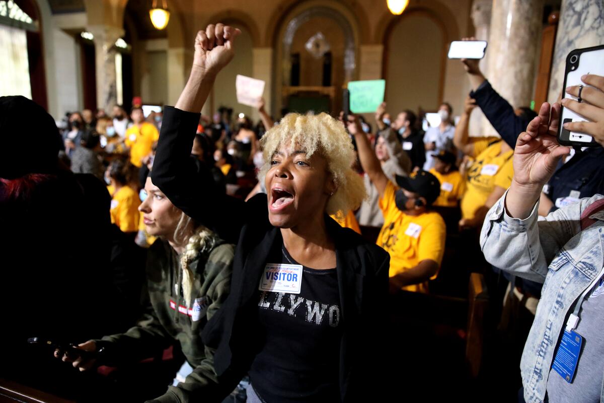 A woman among a crowd inside a city building holds up a clenched fist and calls out.