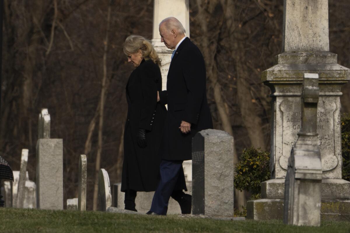 President Biden and First Lady Jill Biden walk between tombstones to attend Mass at a church.