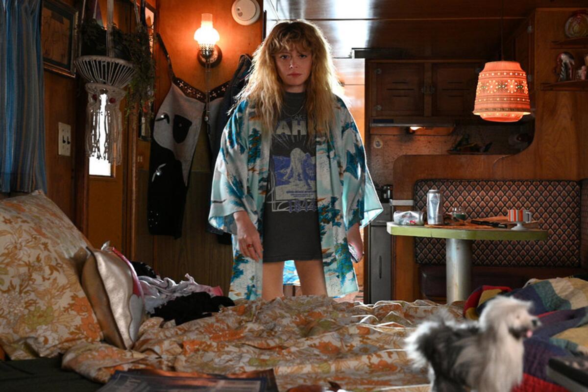 A woman in a T-shirt and open robe stands in a cramped and messy bedroom.