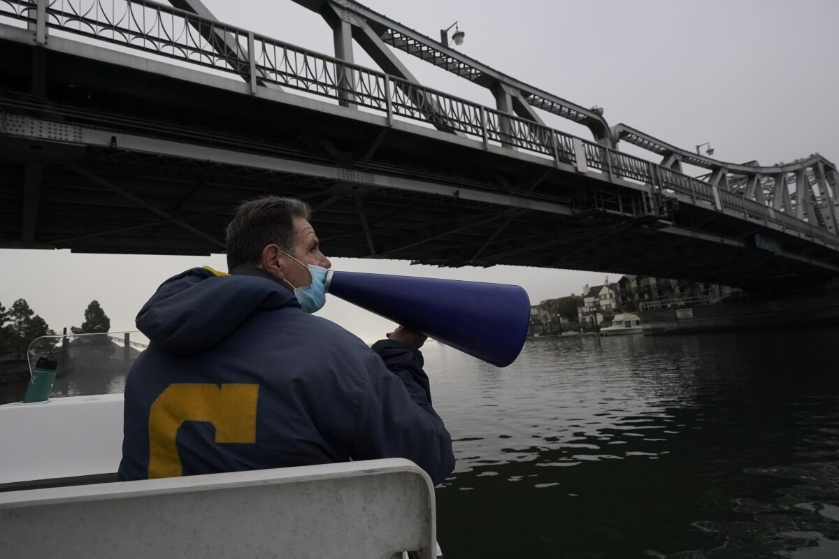Coach Mike Teti speaks into a megaphone toward rowers practicing in the Oakland Estuary in Oakland, Calif., Thursday, Nov. 19, 2020. Nearly two dozen U.S. men's rowers signed on to keep training for another year after the Tokyo Olympics were delayed until 2021. Teti, a three-time Olympian himself, is thrilled the group stuck together. (AP Photo/Jeff Chiu)