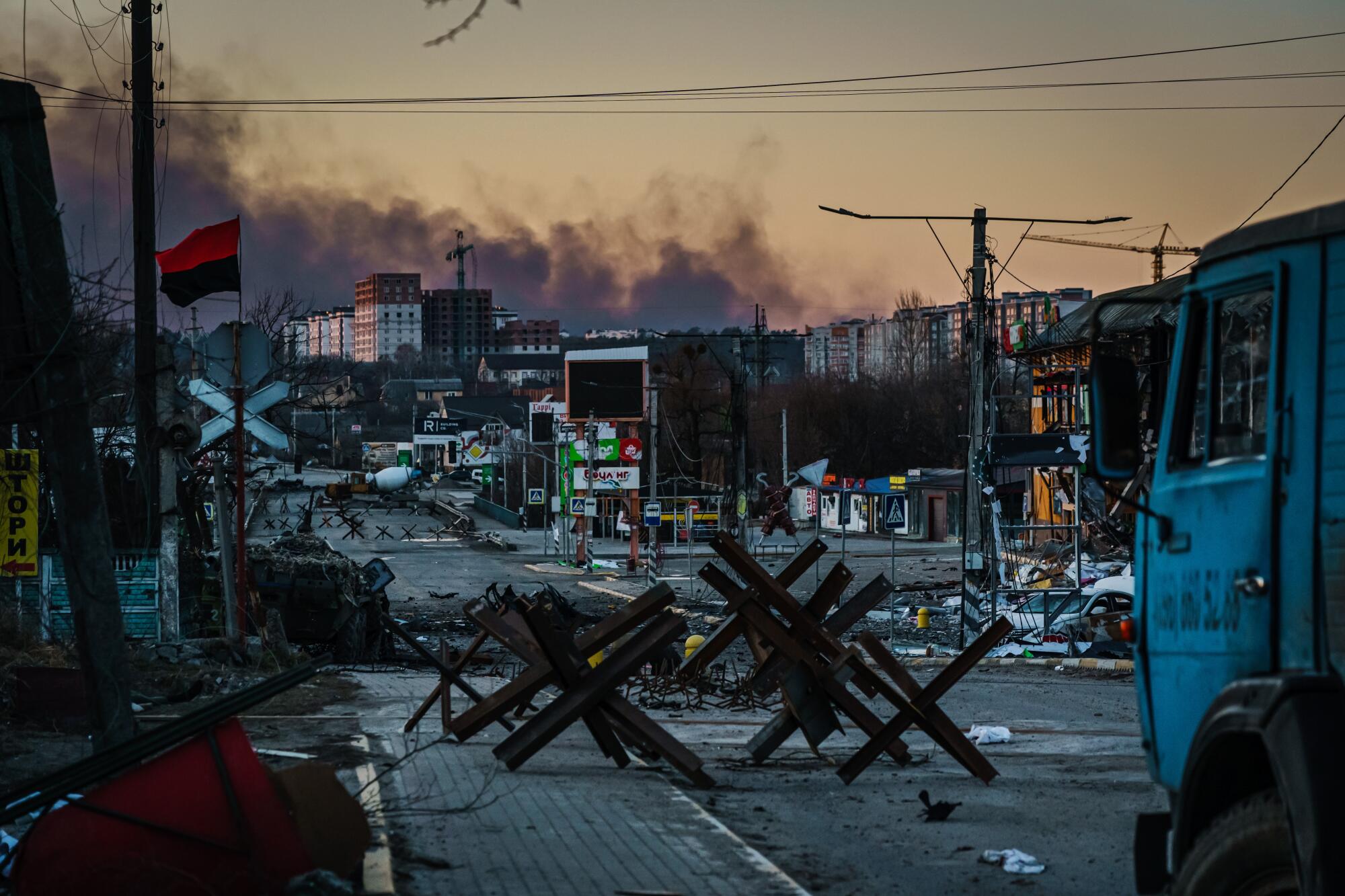 A street with obstacles across it in several places while smoke rises behind buildings.