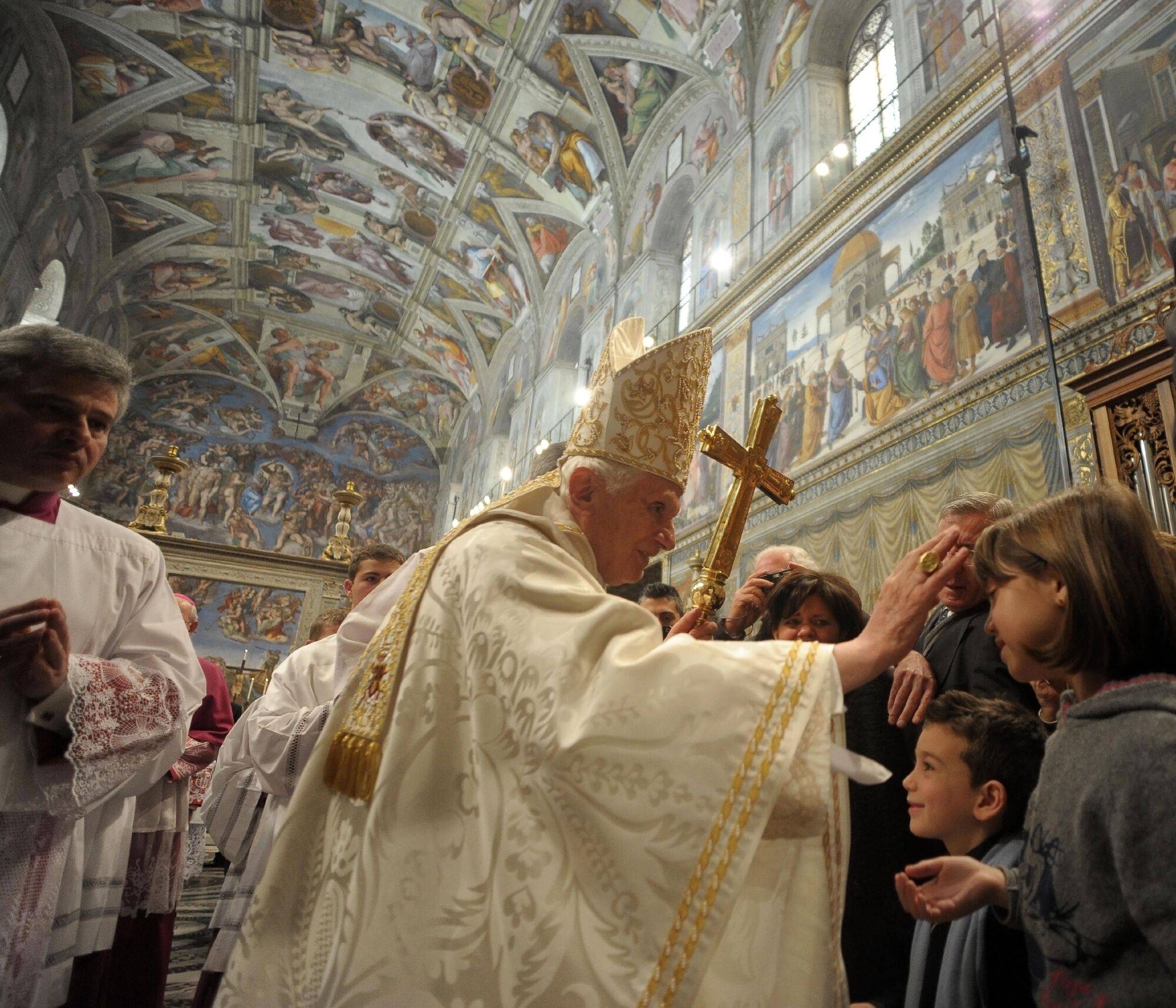 Pope Benedict XVI  in cream-colored robes shakes hands with children in the Sistine Chapel.