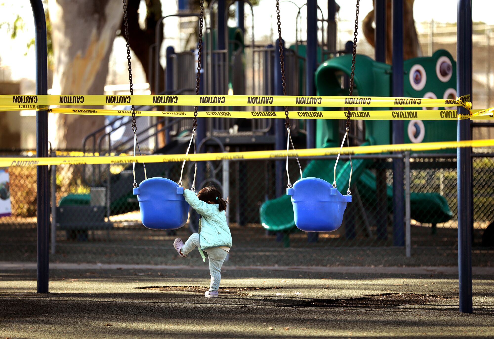 A toddler tries to climb onto a swing at a playground surrounded by yellow caution tape.