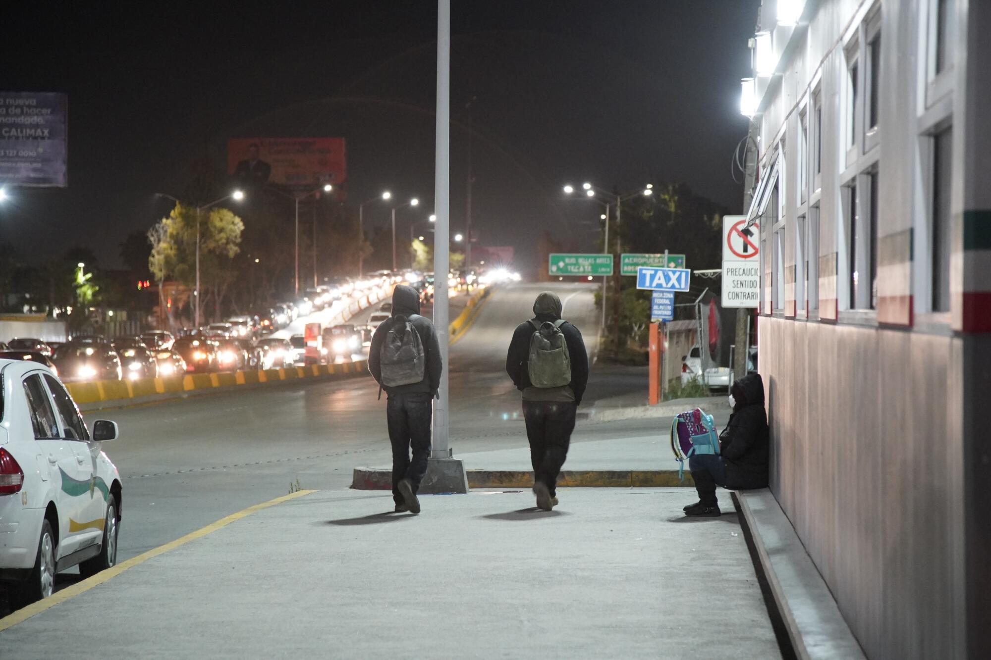 Two men wearing backpacks and hoodies walk out of the Otay Mesa Port of Entry area 