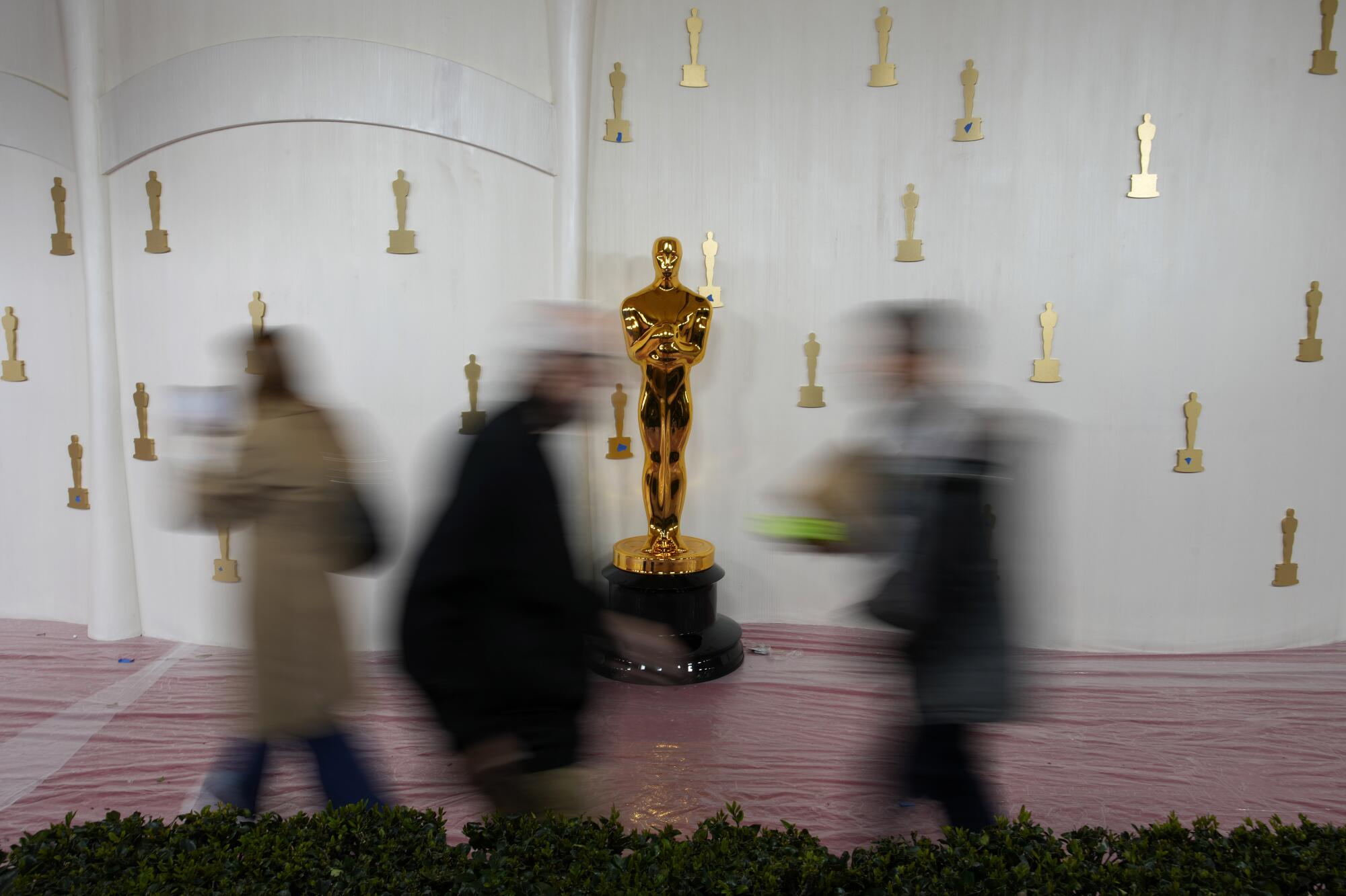 A golden Oscars statuette stands against a white wall with three blurry profiles of people passing in front of it.