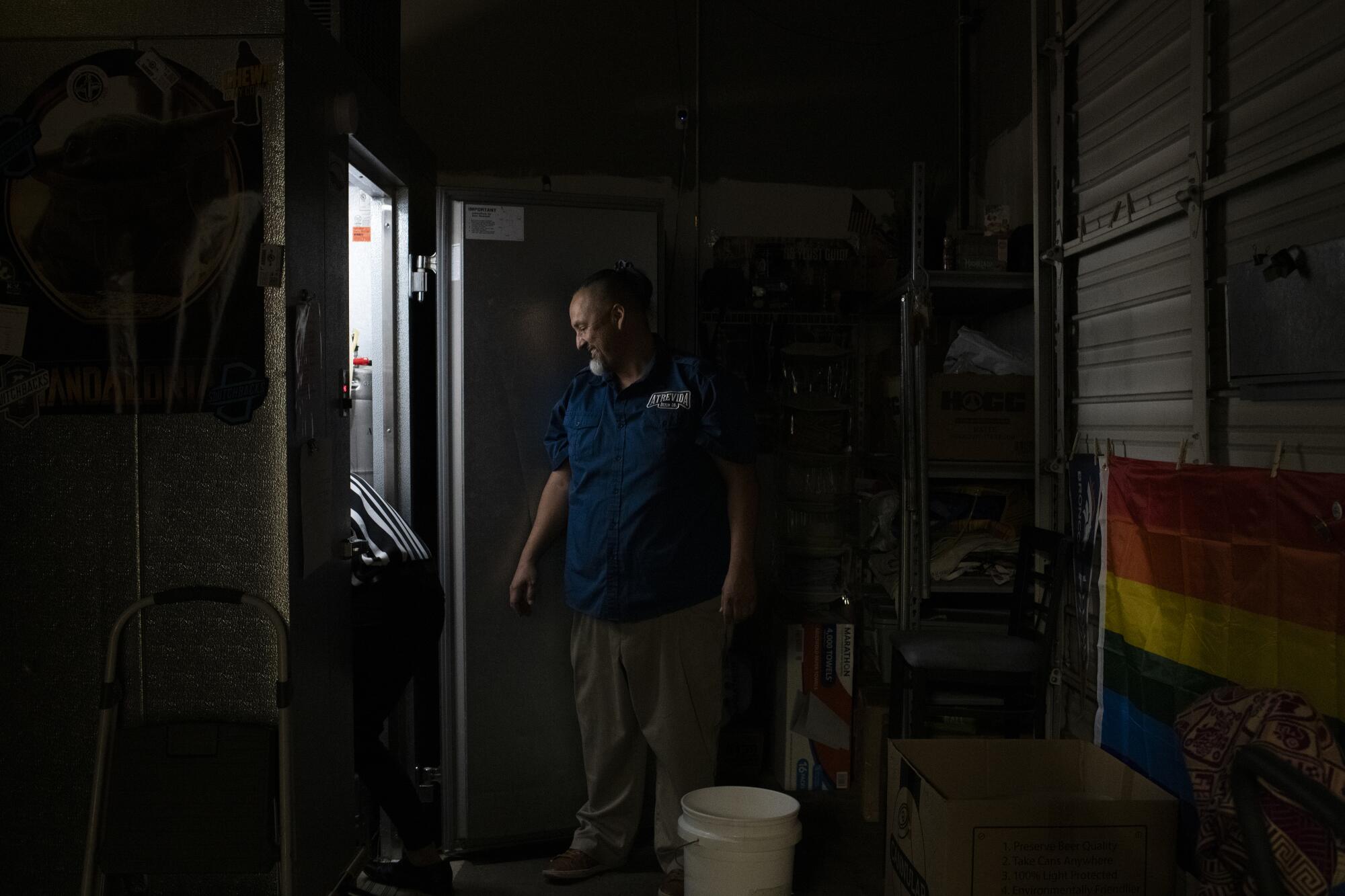 A man in a blue shirt and khaki pants stands look through a doorway, a bucket near his feet