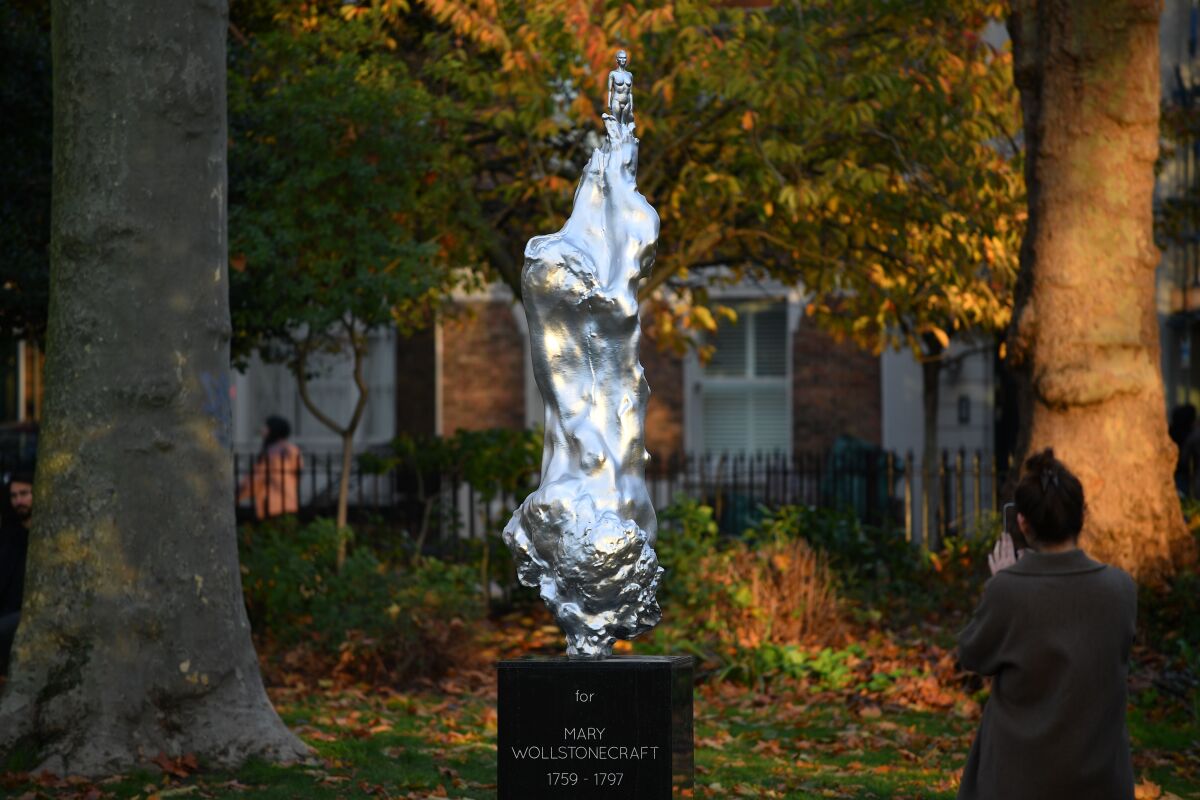 A wide view of a bright silver sculpture of Mary Wollstonecraft in a leafy London park