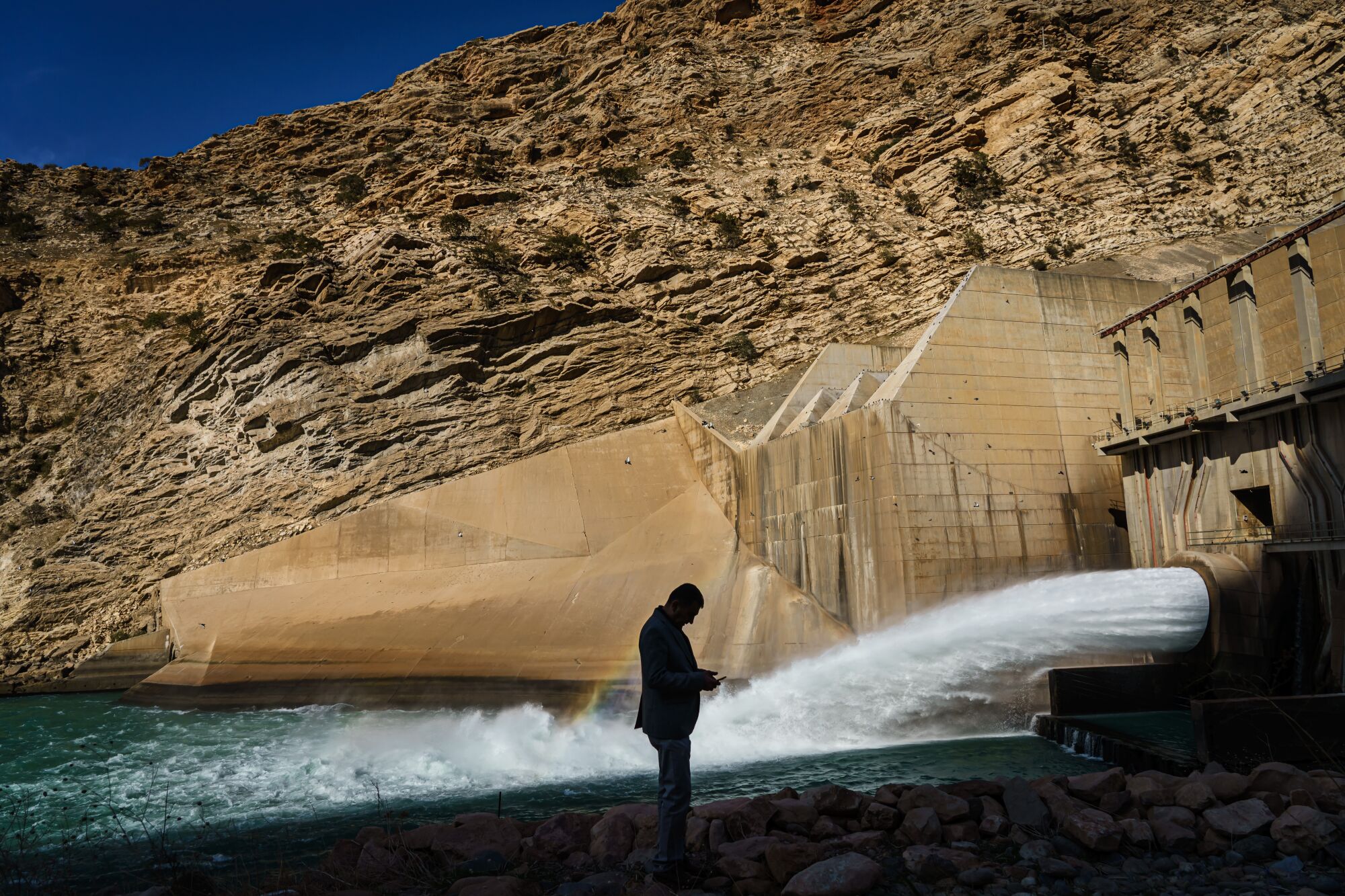 A silhouette of a man standing near where water is gushing out of a pipe, right, at a dam 
