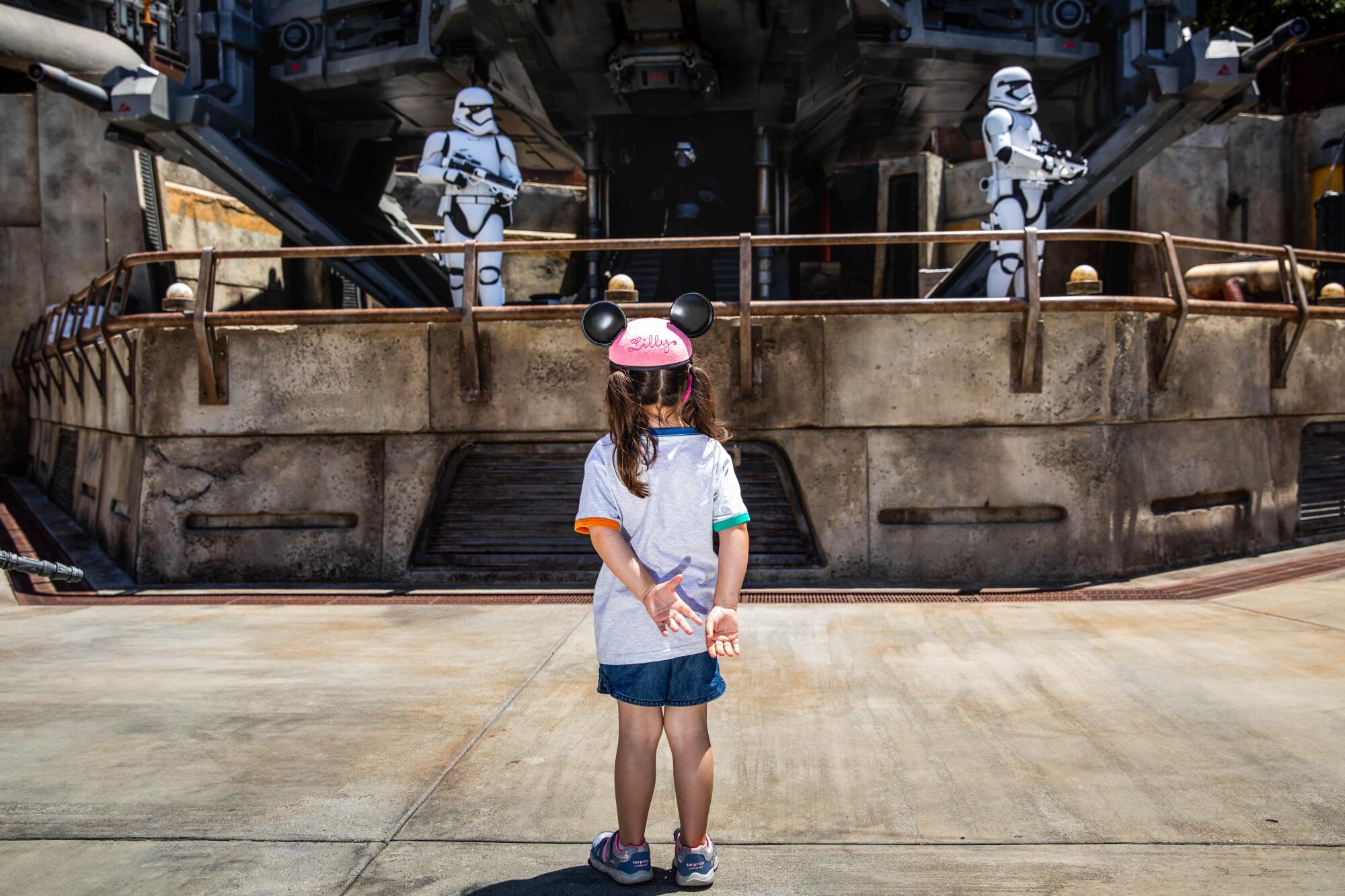 A girl in a pink mouse-ears hat looks at two performers on a Star Wars set in helmeted costumes, holding fake weapons.