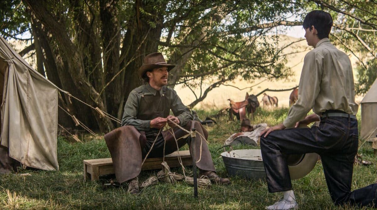 A man in a cowboy hat sitting under a tree talks to a younger man.