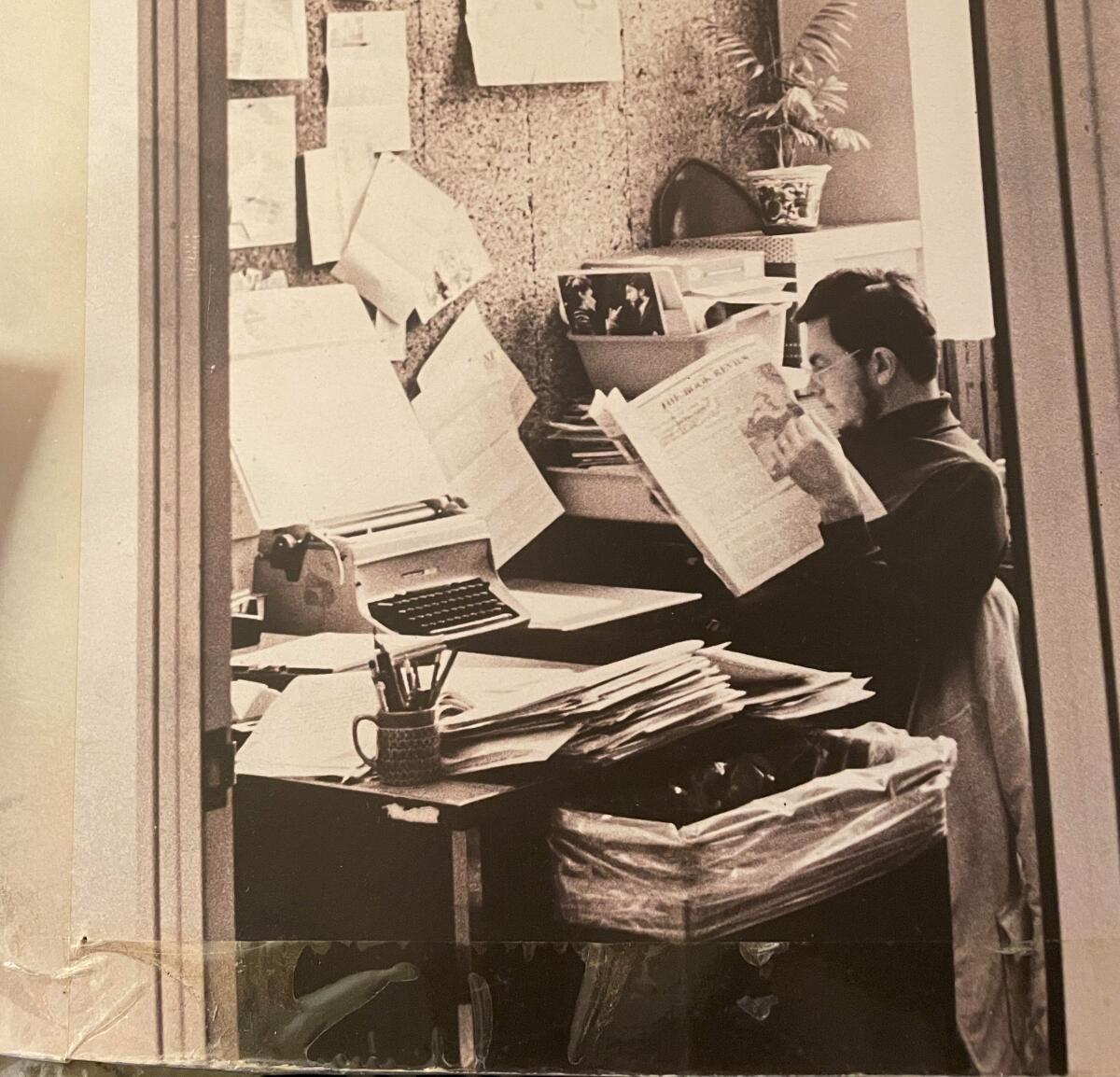 A man sits at a work desk reading a newspaper.